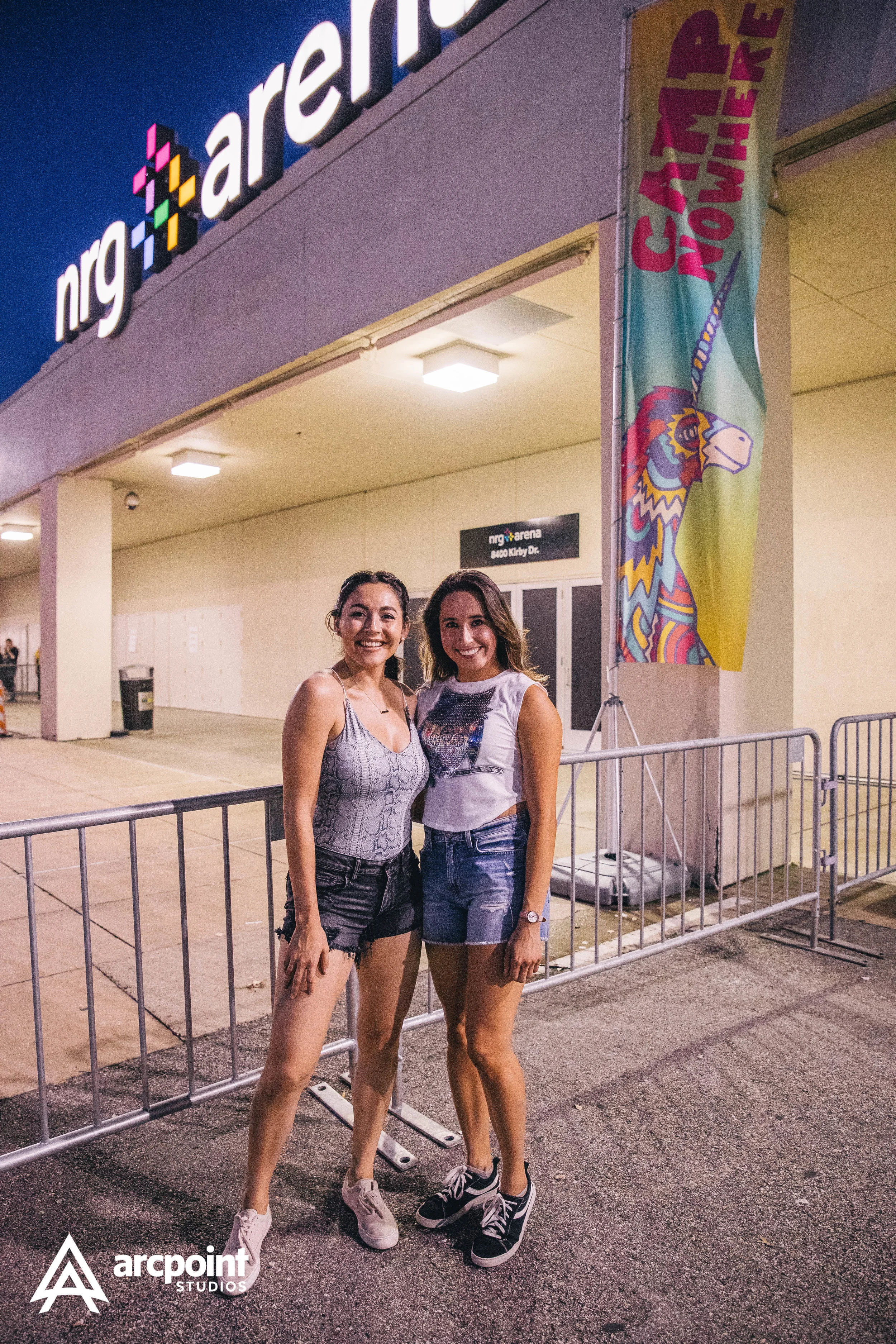Two young women standing outside the entrance of an arena, smiling at the camera, at night. There is a colorful flag with 