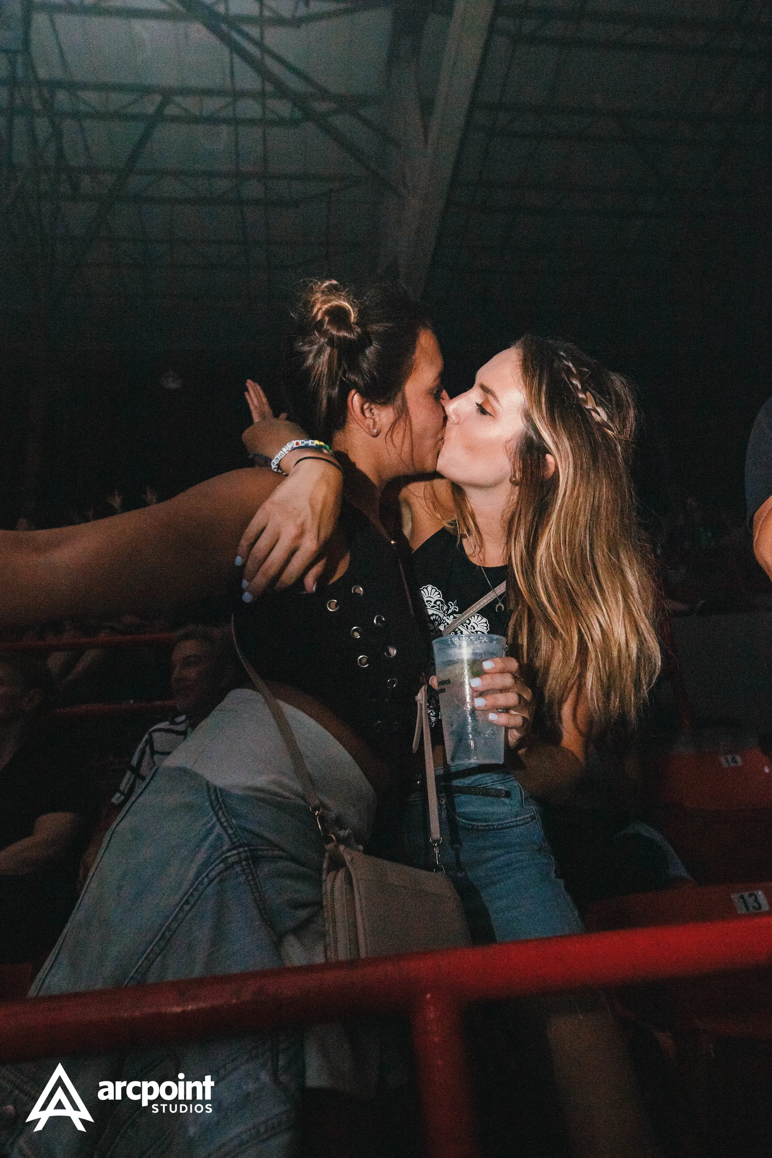 Two women sharing a kiss at a concert or event with dark lighting and a metal structure overhead.