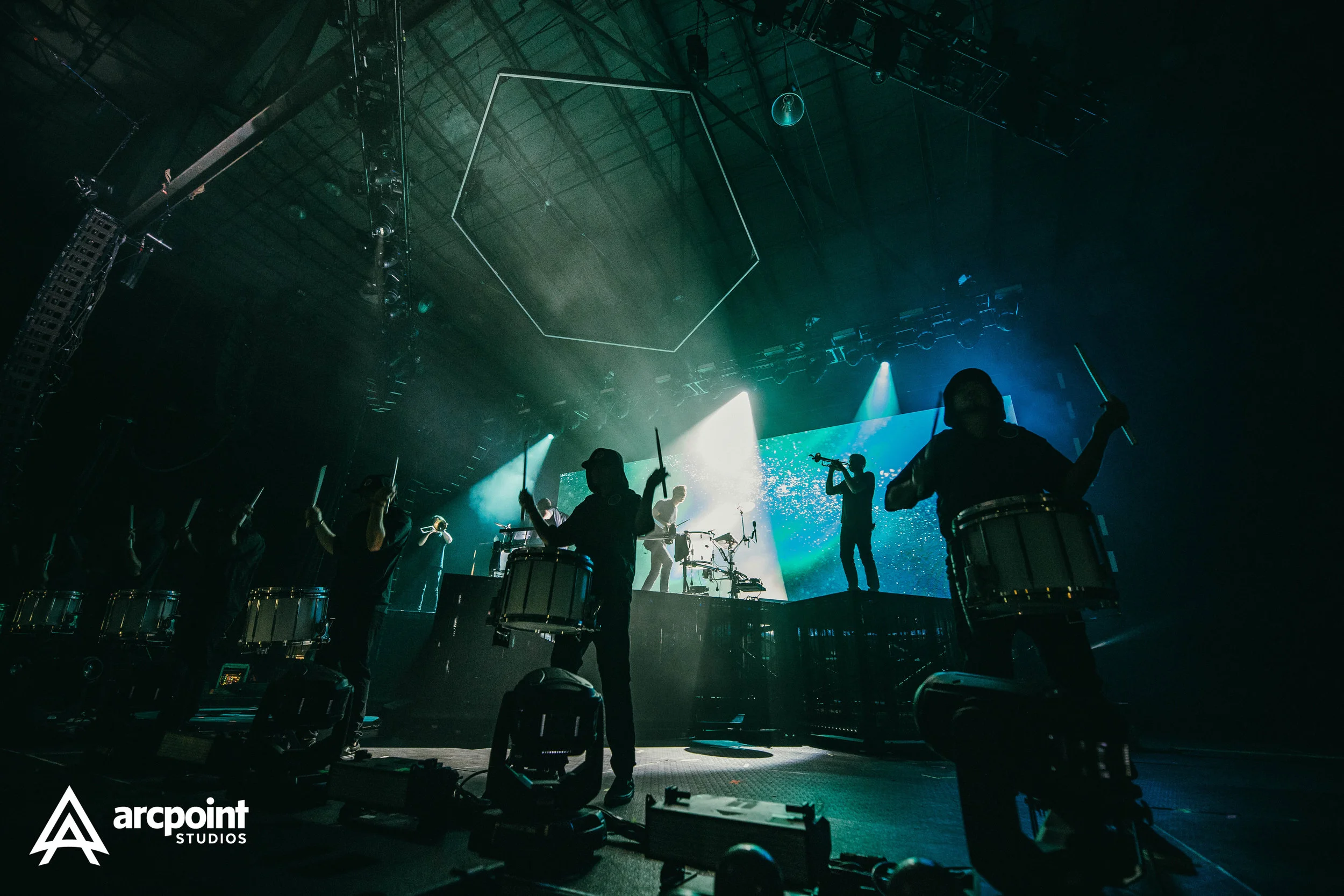 A live concert performance with musicians playing drums, flutes, and other instruments on stage, illuminated by blue and green lighting with stage equipment visible.