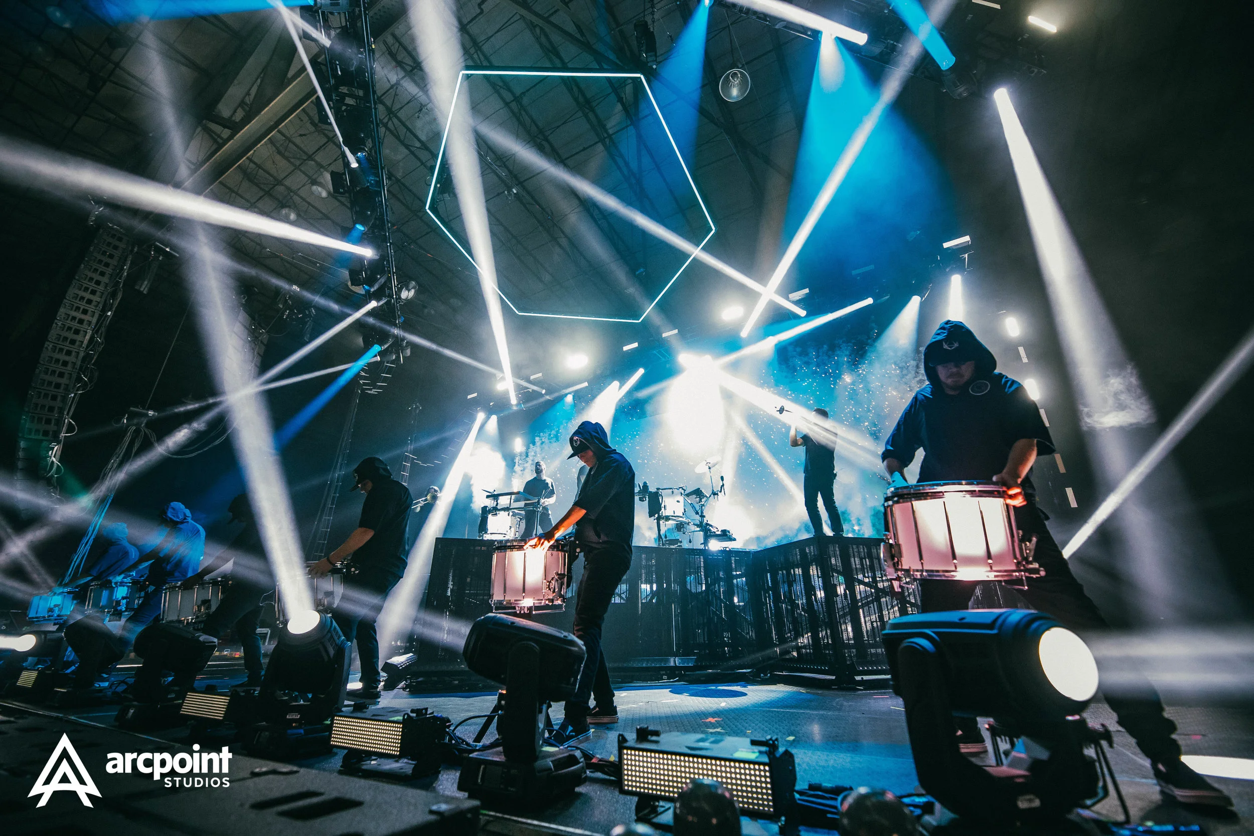 A live music performance with a band on stage, illuminated by colorful stage lights and laser effects, with musicians playing instruments under a dark, industrial-style ceiling.