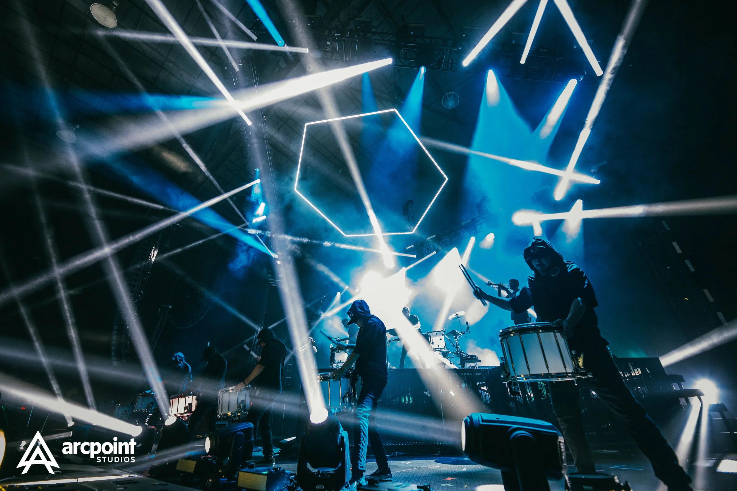 A live music performance with musicians playing drums and other instruments on stage, surrounded by bright white and blue stage lights and geometric light fixtures overhead.