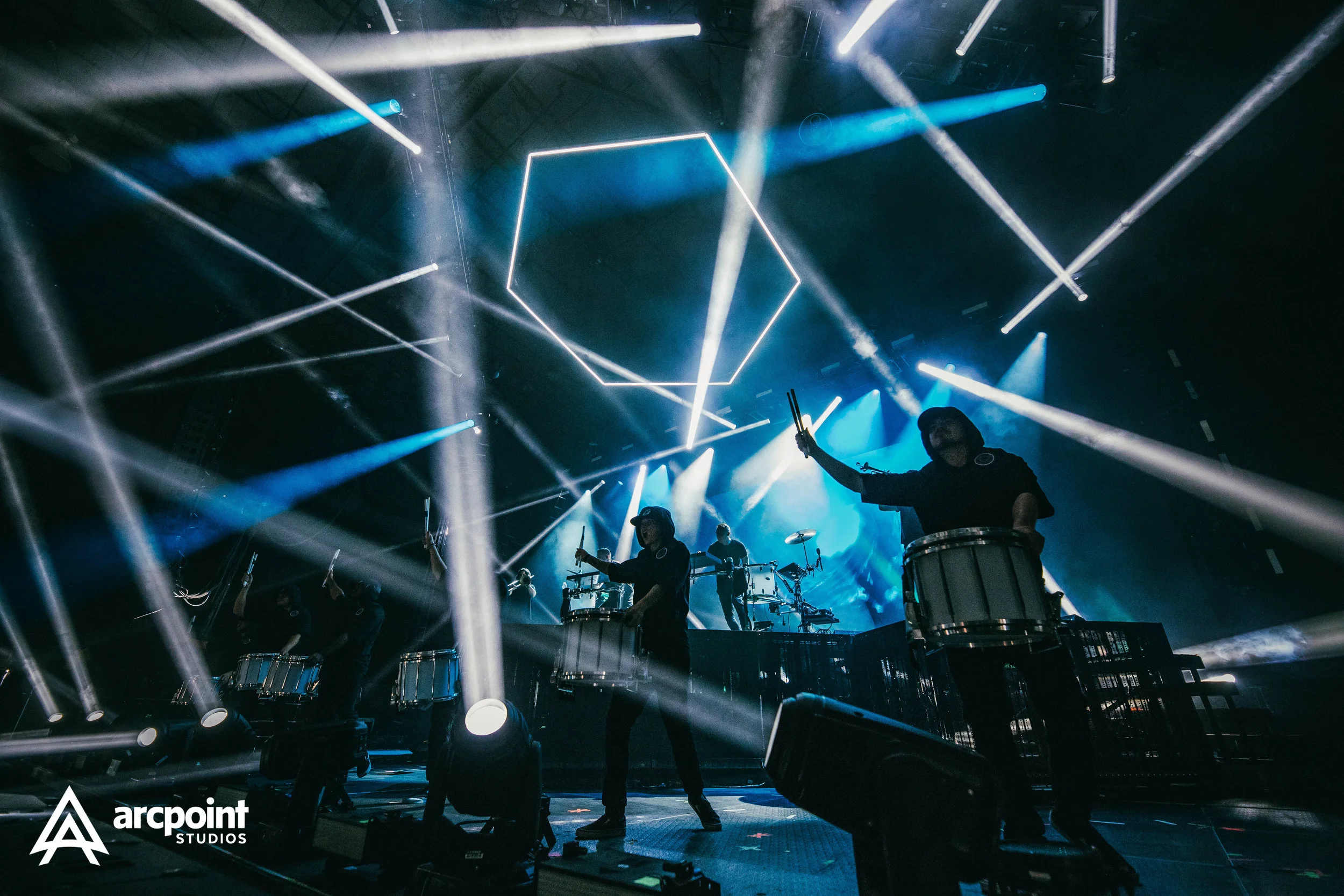 A live concert performance with musicians on stage, illuminated by dynamic blue and white stage lights, featuring drums, digital equipment, and a large geometric light fixture hanging above.