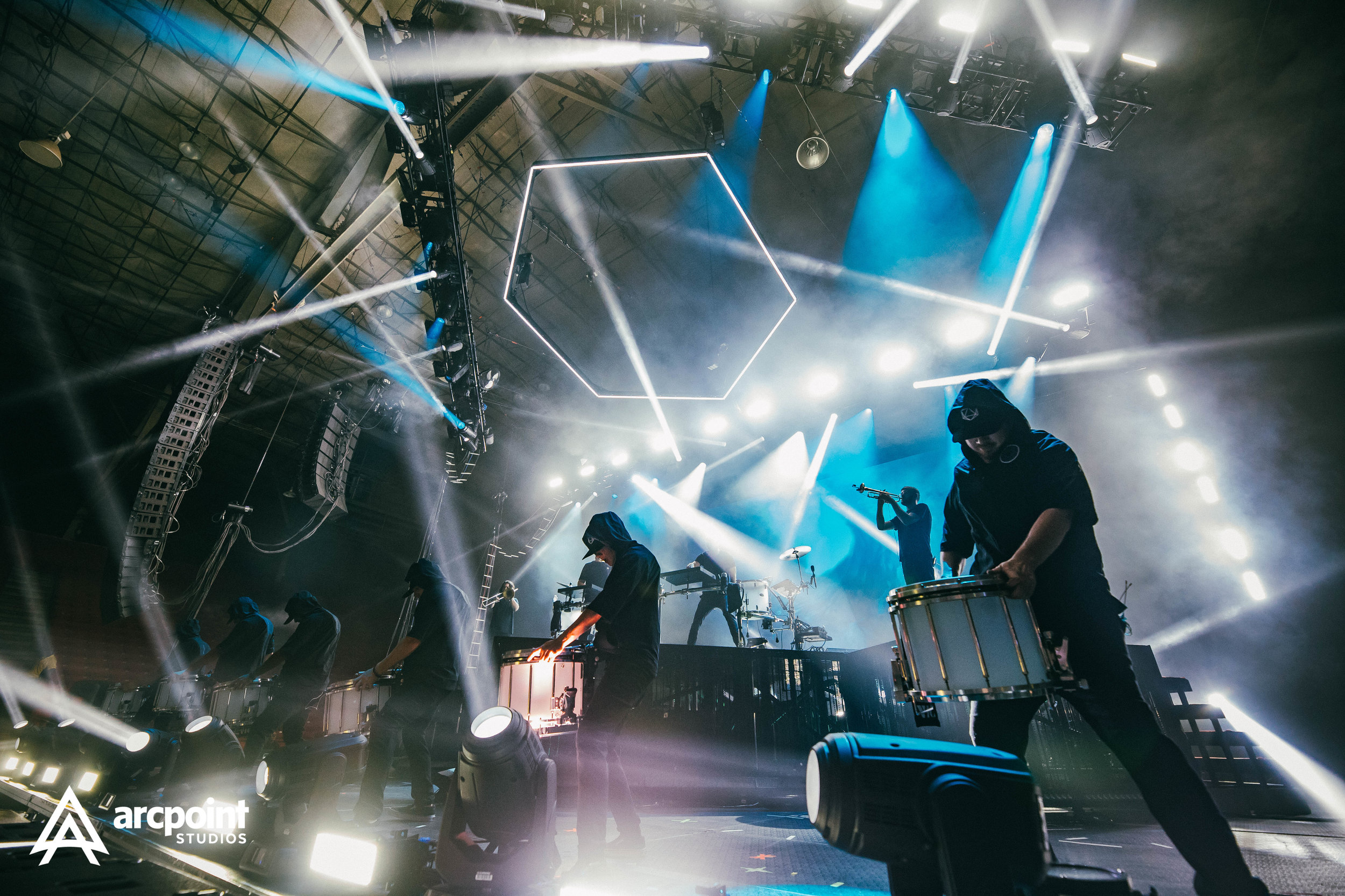 Musicians performing on stage with colorful lights and geometric light fixtures, some playing drums, trumpet, and other instruments, in a concert setting.