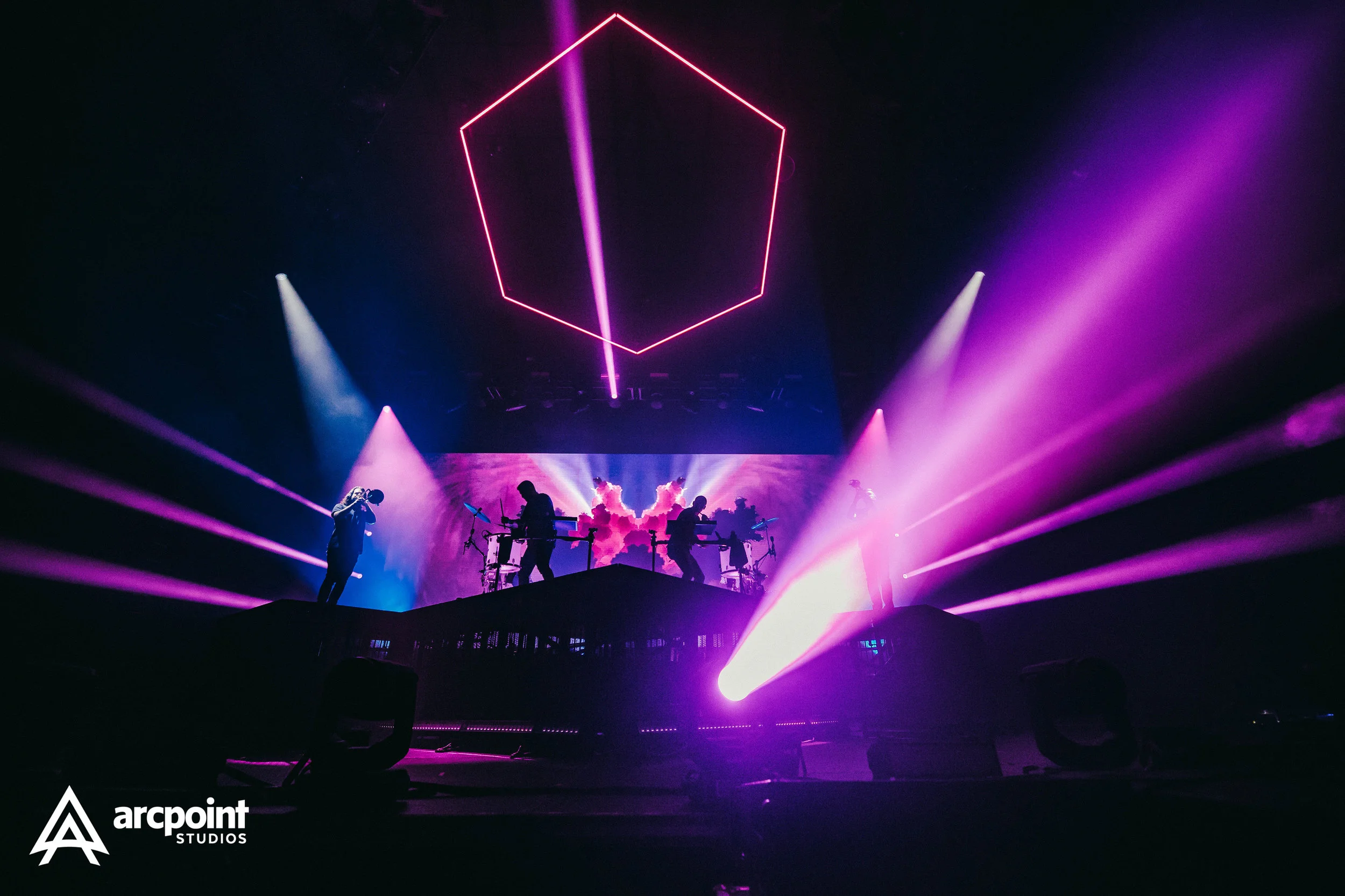 A live concert stage with vibrant purple and pink lighting, a digital hexagon shape with a line through it projected above, silhouettes of musicians playing instruments, and a colorful digital background. Branding for arcpoint STUDIOS is visible in t