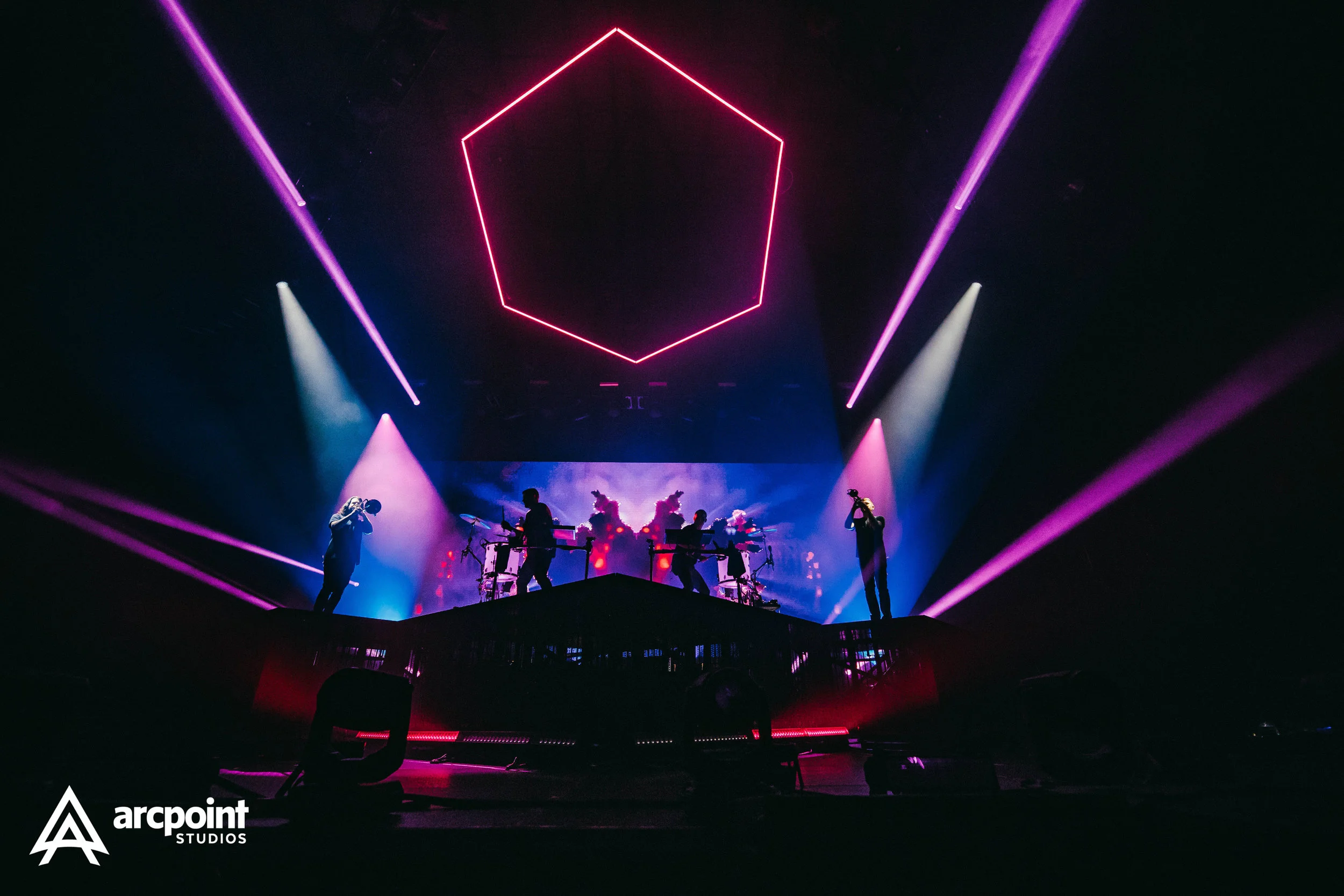 A live concert stage with five musicians, colorful lights, and a neon hexagon above them.