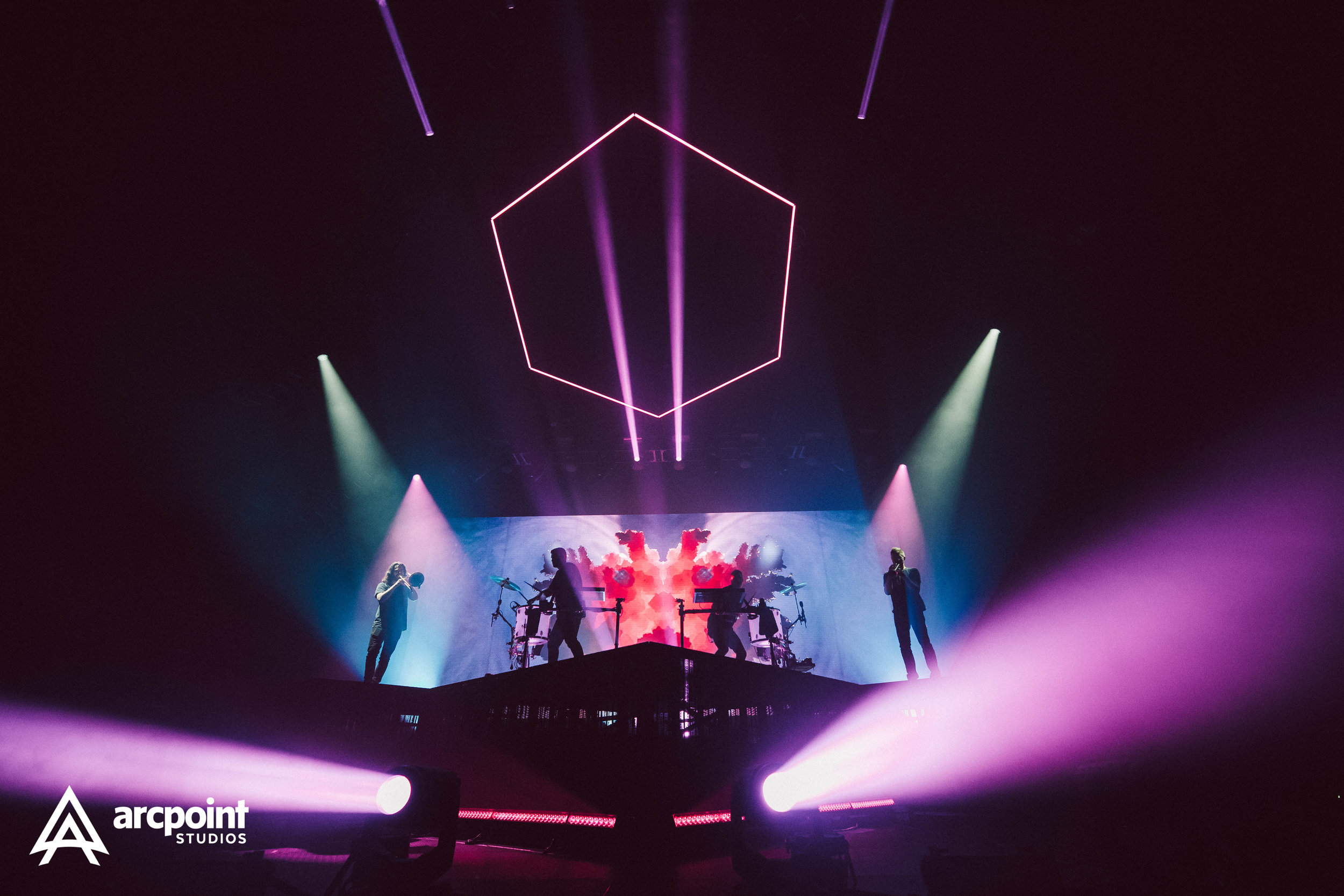 A live concert performance on a stage with colorful lights, silhouettes of musicians, a large screen with abstract visuals, and a geometric neon light above.