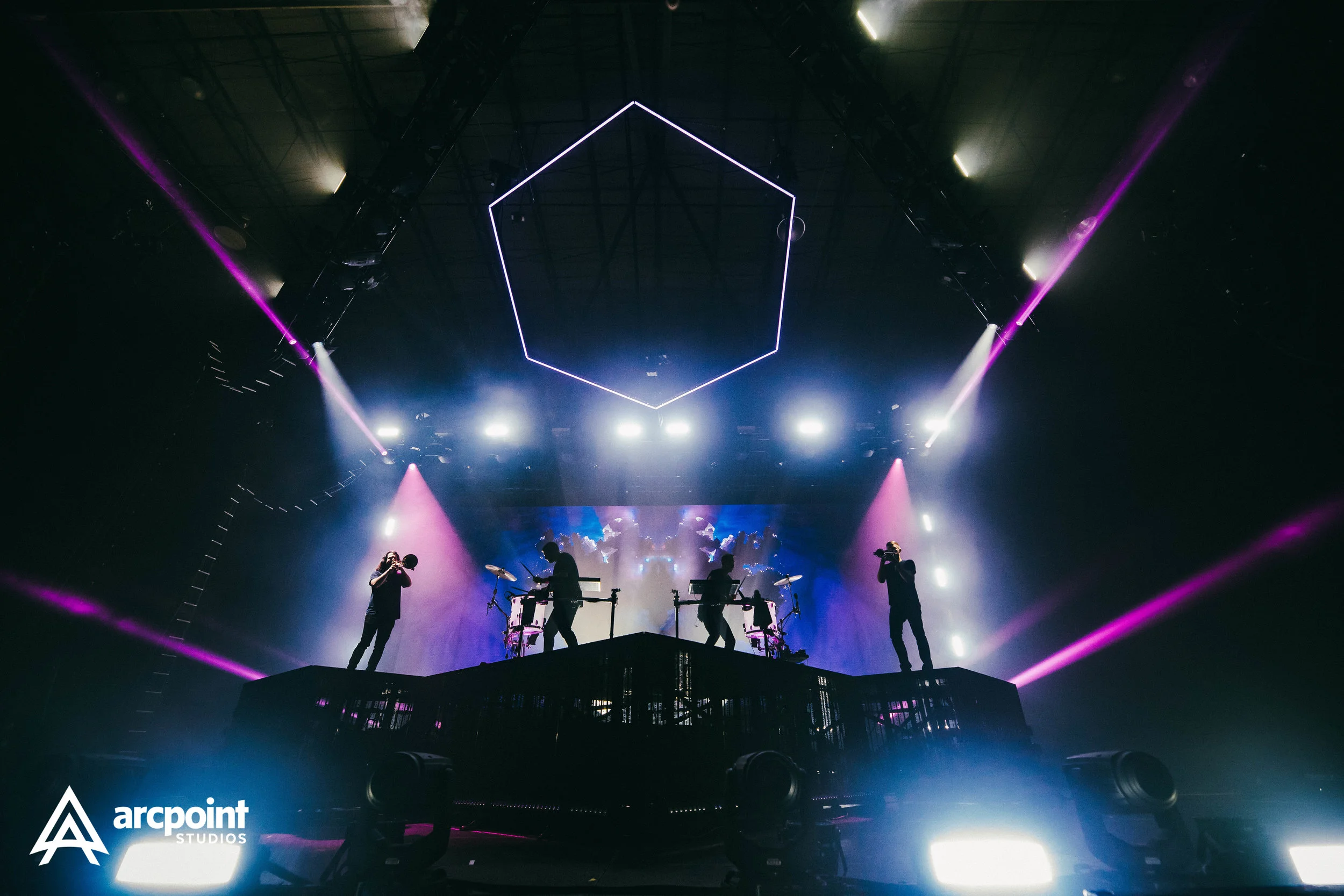 Silhouettes of four musicians performing on a stage with bright lights and purple laser beams in a dark indoor venue.