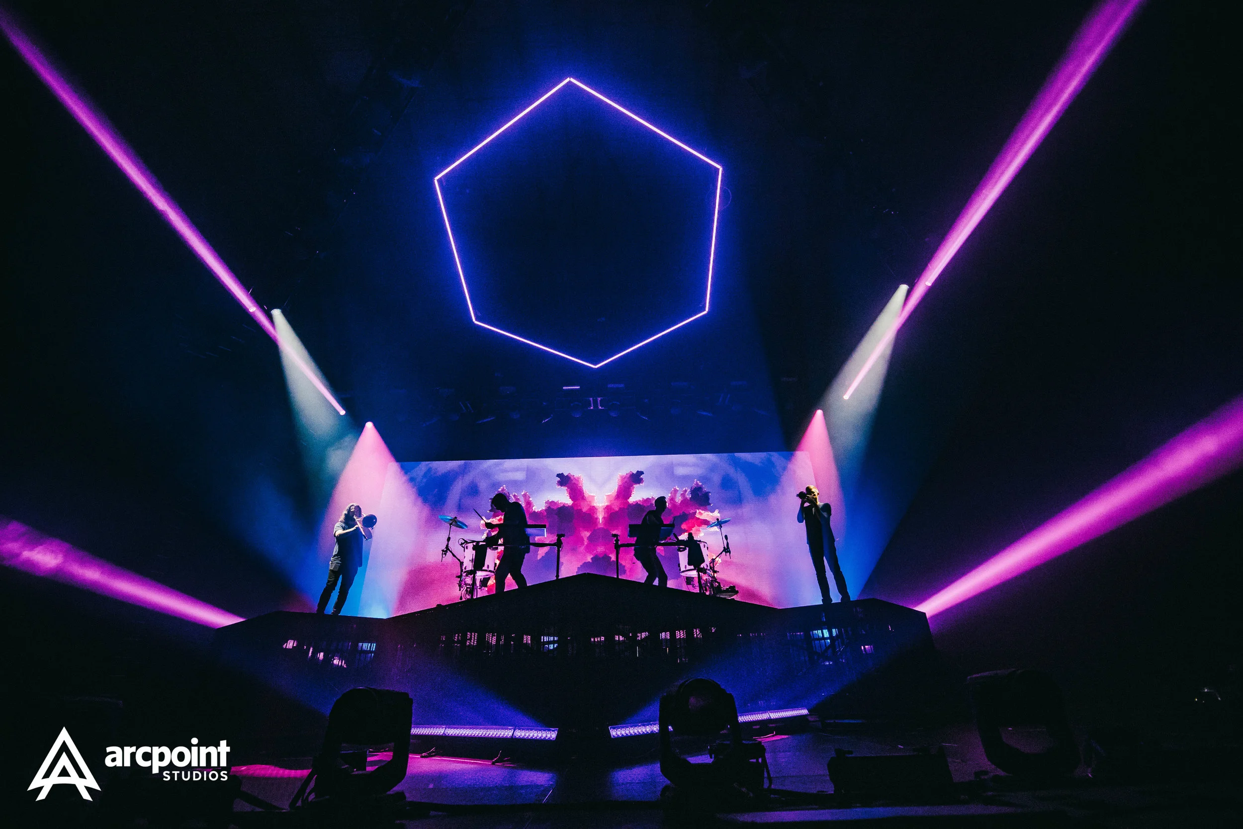 A live concert stage with musicians and visual effects. There are purple and blue lighting, a large hexagon neon light in the ceiling, and a screen displaying pink and purple clouds. The band's logo 'arcpoint Studios' is in the bottom left corner.