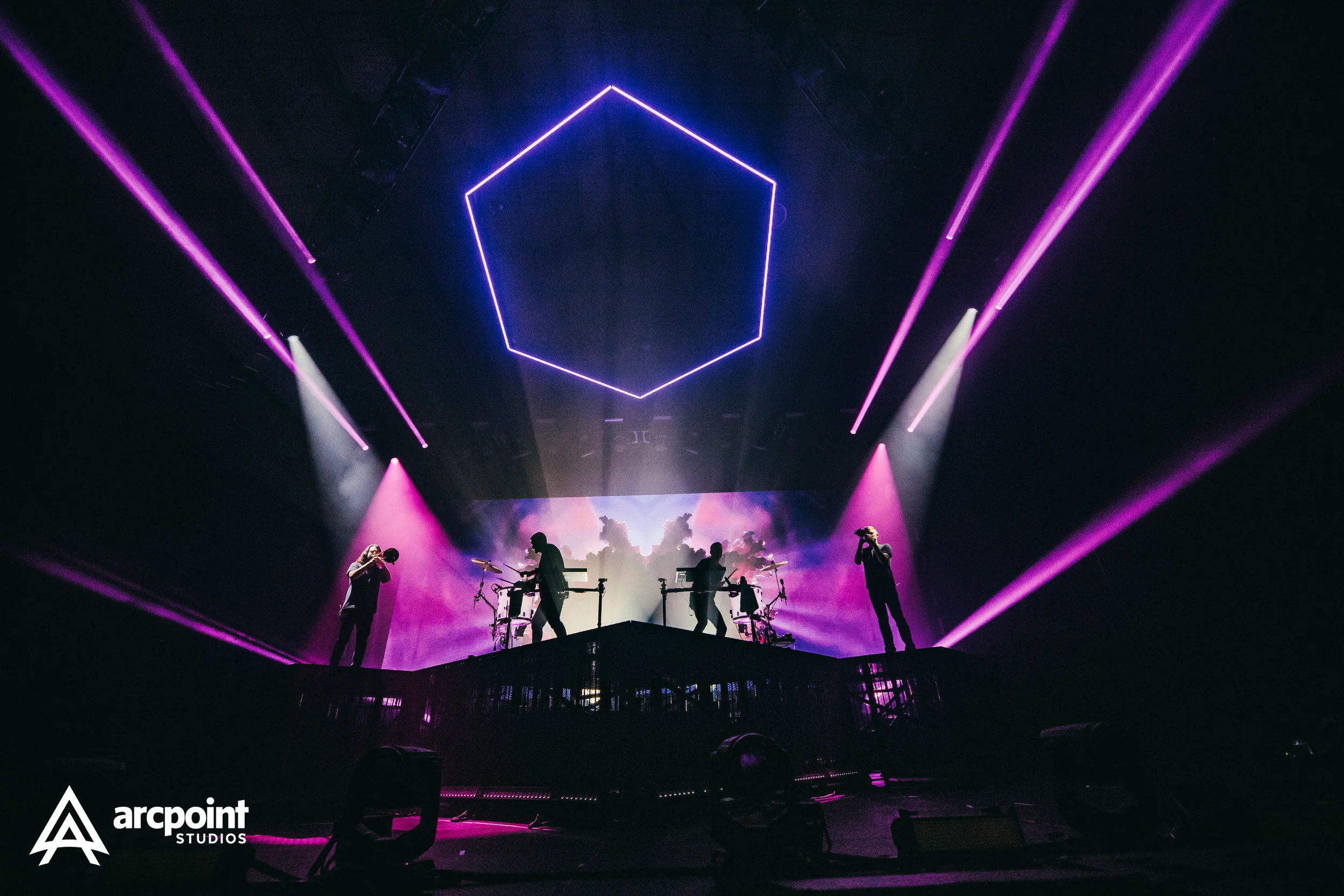 Concert stage with three musicians performing amid purple and pink stage lights, large projection screen with a colorful cloud scene in the background, and geometric neon shape overhead.