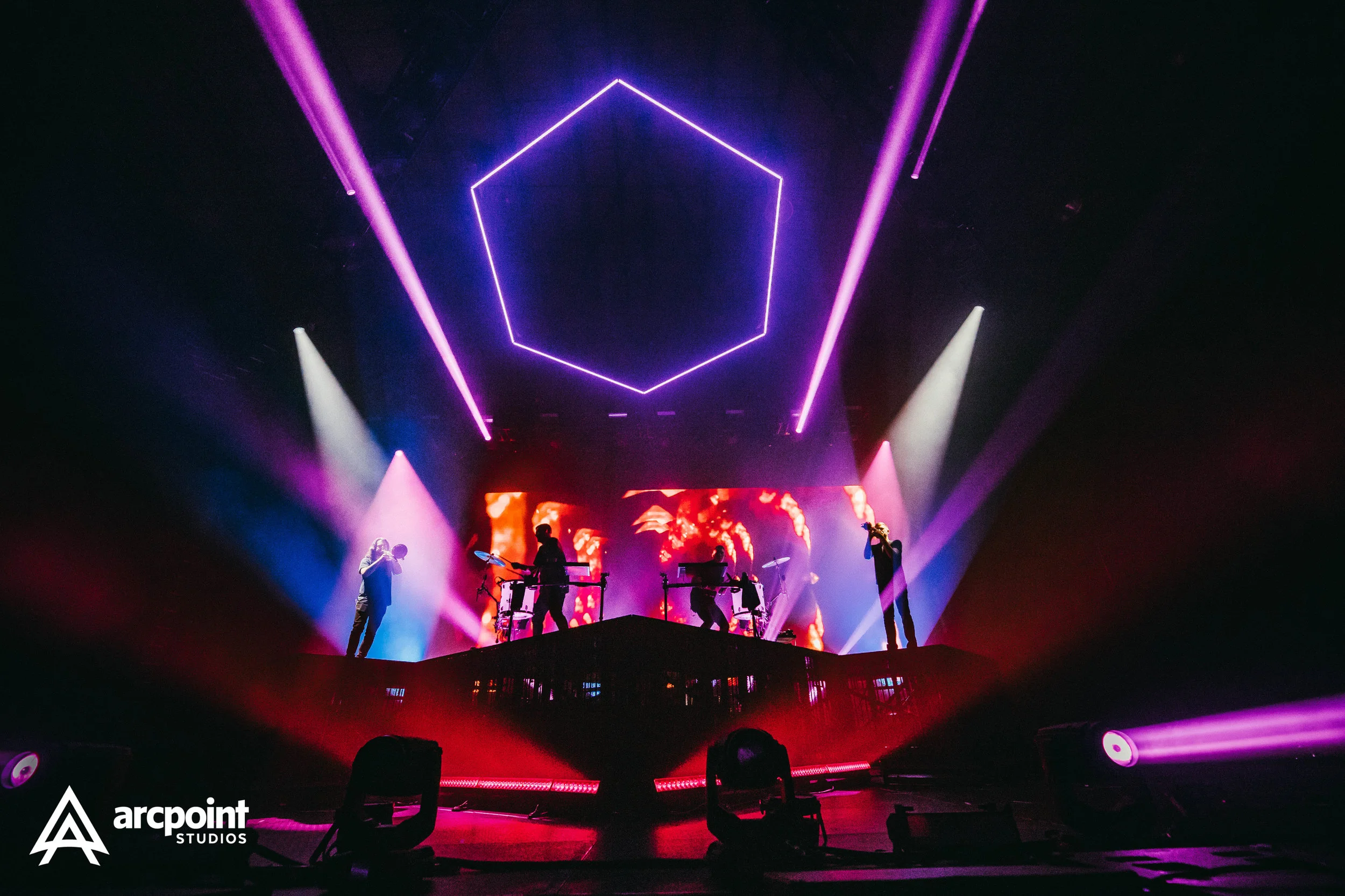 A live concert performance on a stage with colorful lights, a large hexagon-shaped neon light overhead, and musicians playing instruments. The background features a digital screen with fiery visuals, and the lighting creates an energetic atmosphere.