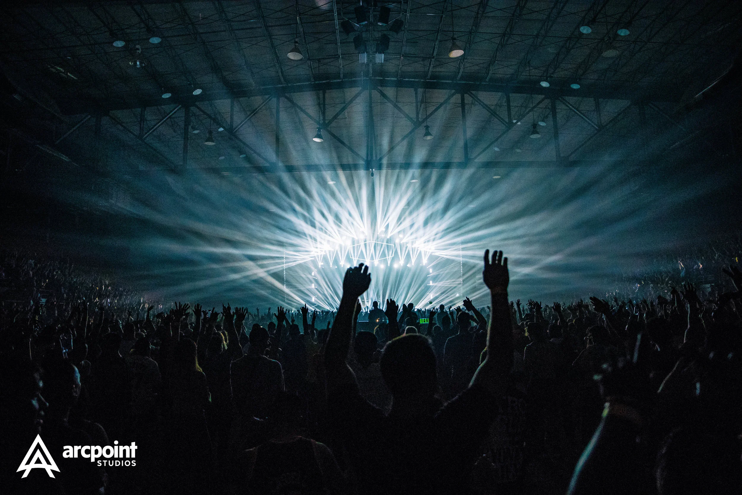 Crowd at a concert with hands raised, stage lights illuminating the venue, and a DJ or performer visible in the background, inside an indoor arena.