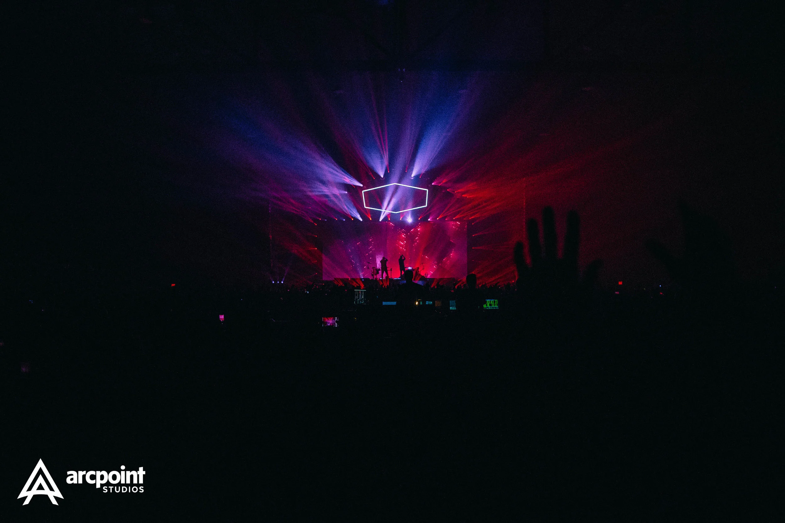 Concert stage with colorful laser lights and silhouettes of performers, audience with raised hands in the foreground, and logo "arcpoint STUDIOS" in the bottom left corner.