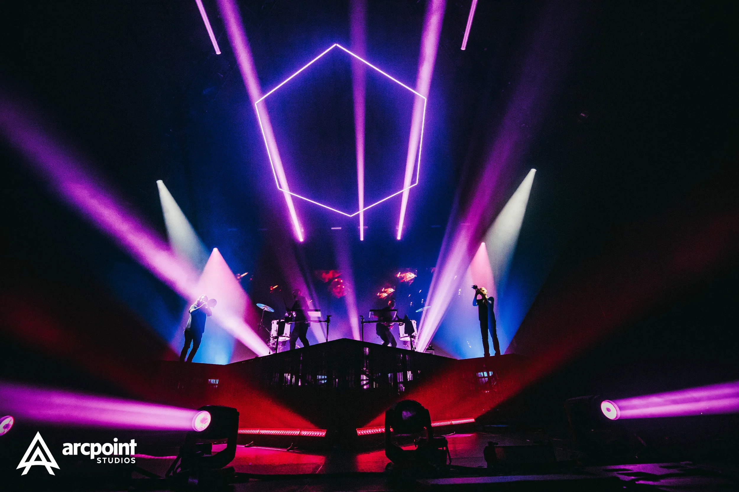A live concert performance with musicians on stage, illuminated by colorful purple, pink, and white lights, with neon geometric shapes overhead, and the Arcpointe Studios logo in the lower left corner.