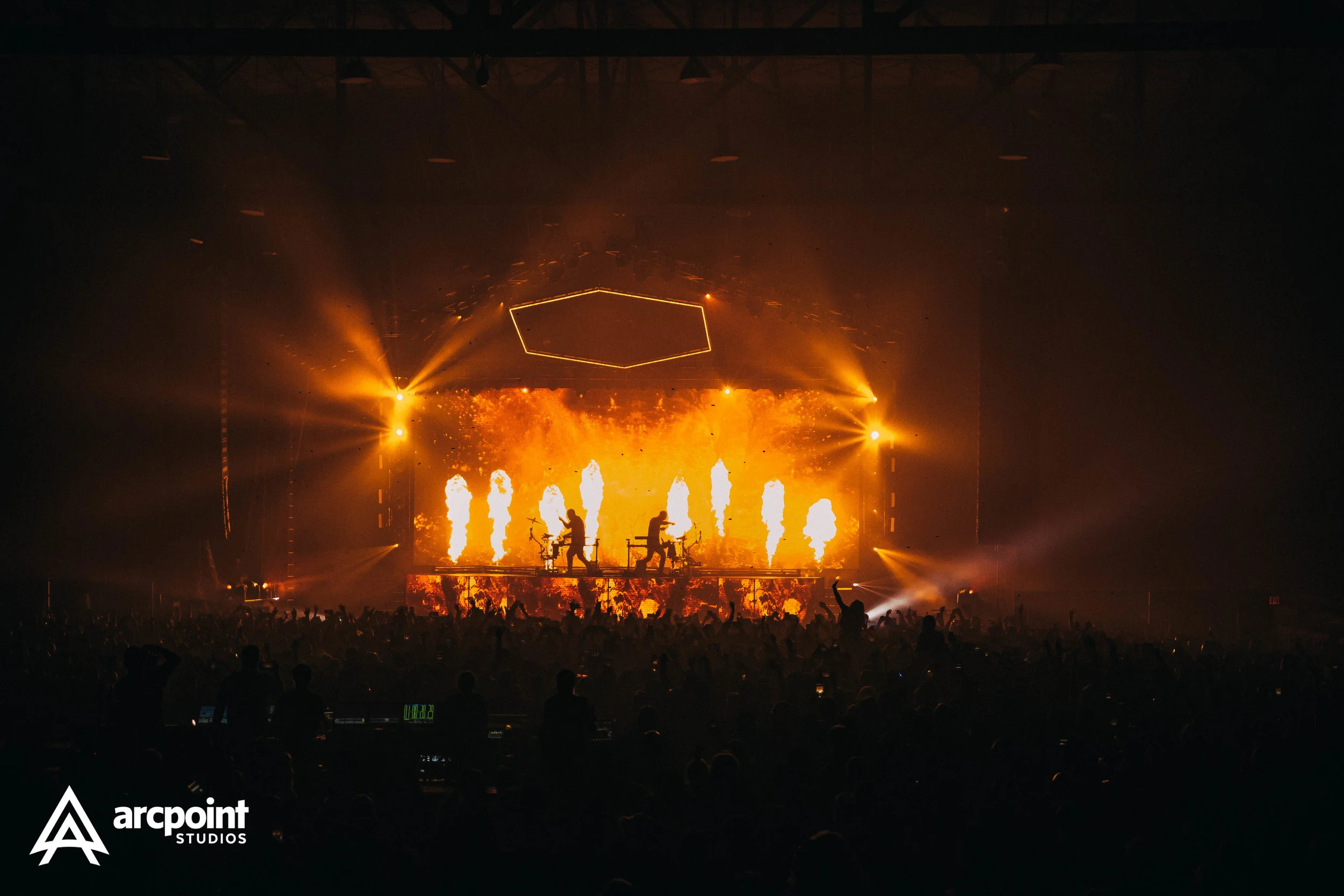 A concert or music festival scene with a large audience, silhouetted in front of a stage with bright orange and yellow flames, pyrotechnics, and dramatic lighting effects, including geometric light shapes hanging above.