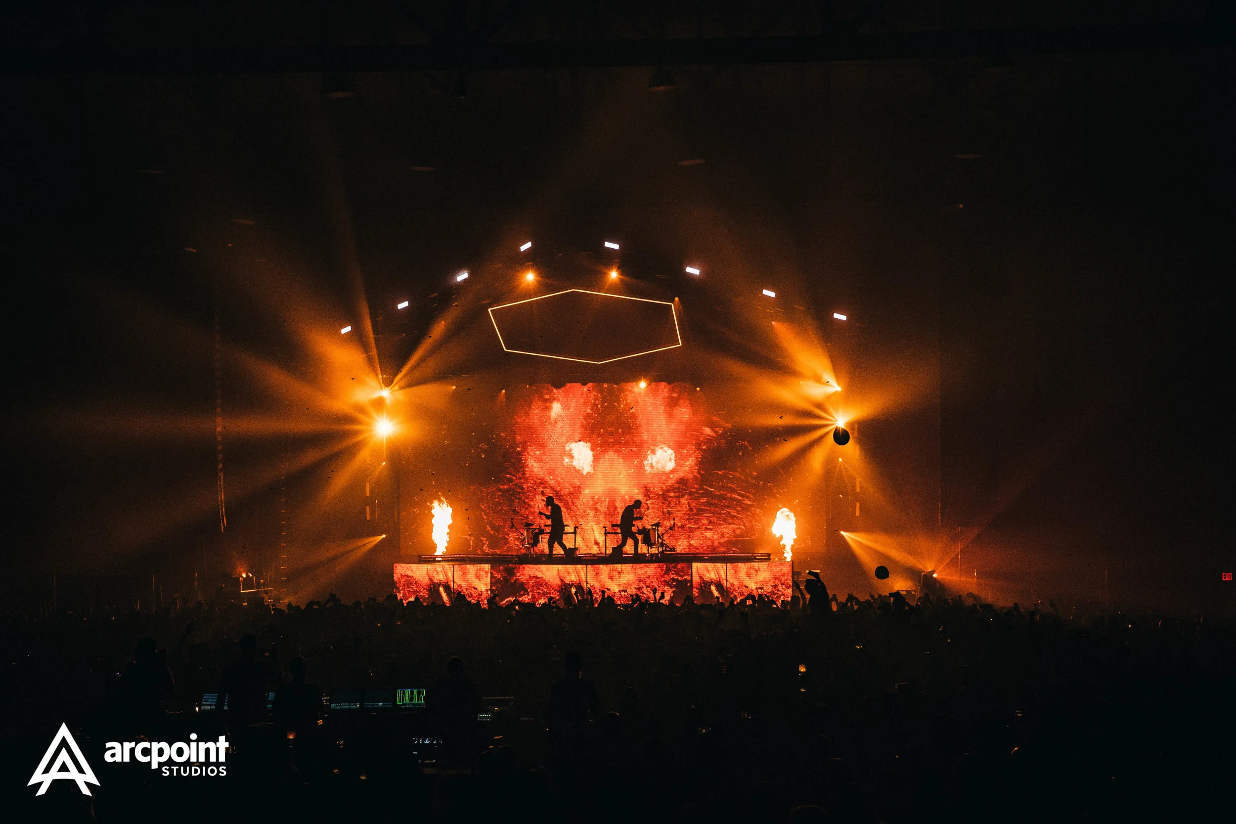 A concert stage with bright orange lighting, two performers on elevated platforms, pyrotechnics, and a large digital screen showing fiery visuals, with an audience in front.