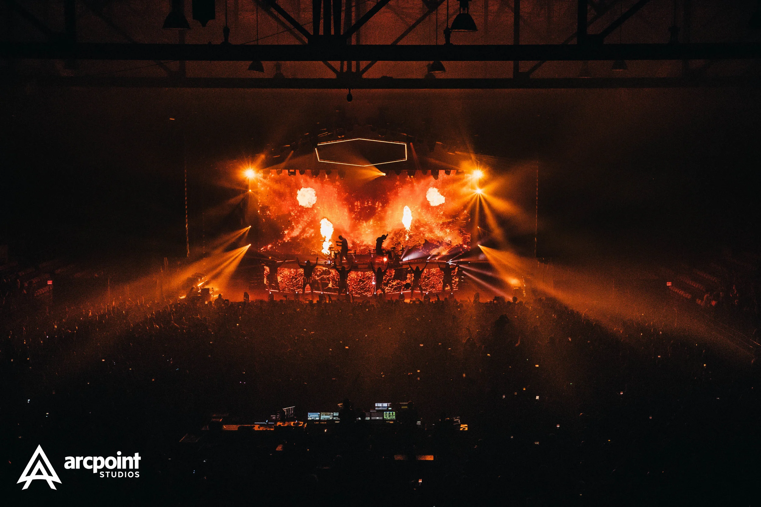 A concert stage with bright orange and yellow lights, fire effects, and performers silhouetted against a large screen displaying fiery visuals, captured in a dark indoor arena filled with audience.