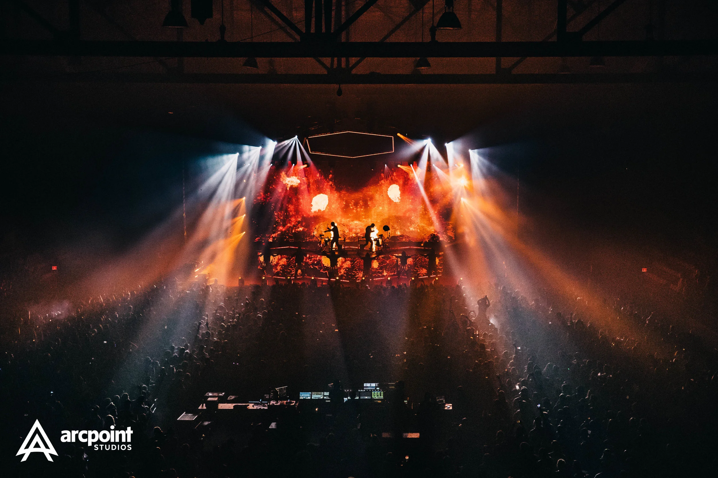A concert stage with bright orange and white lighting effects, a digital background resembling fire, and silhouettes of musicians playing instruments, surrounded by a large audience in a dark indoor venue.