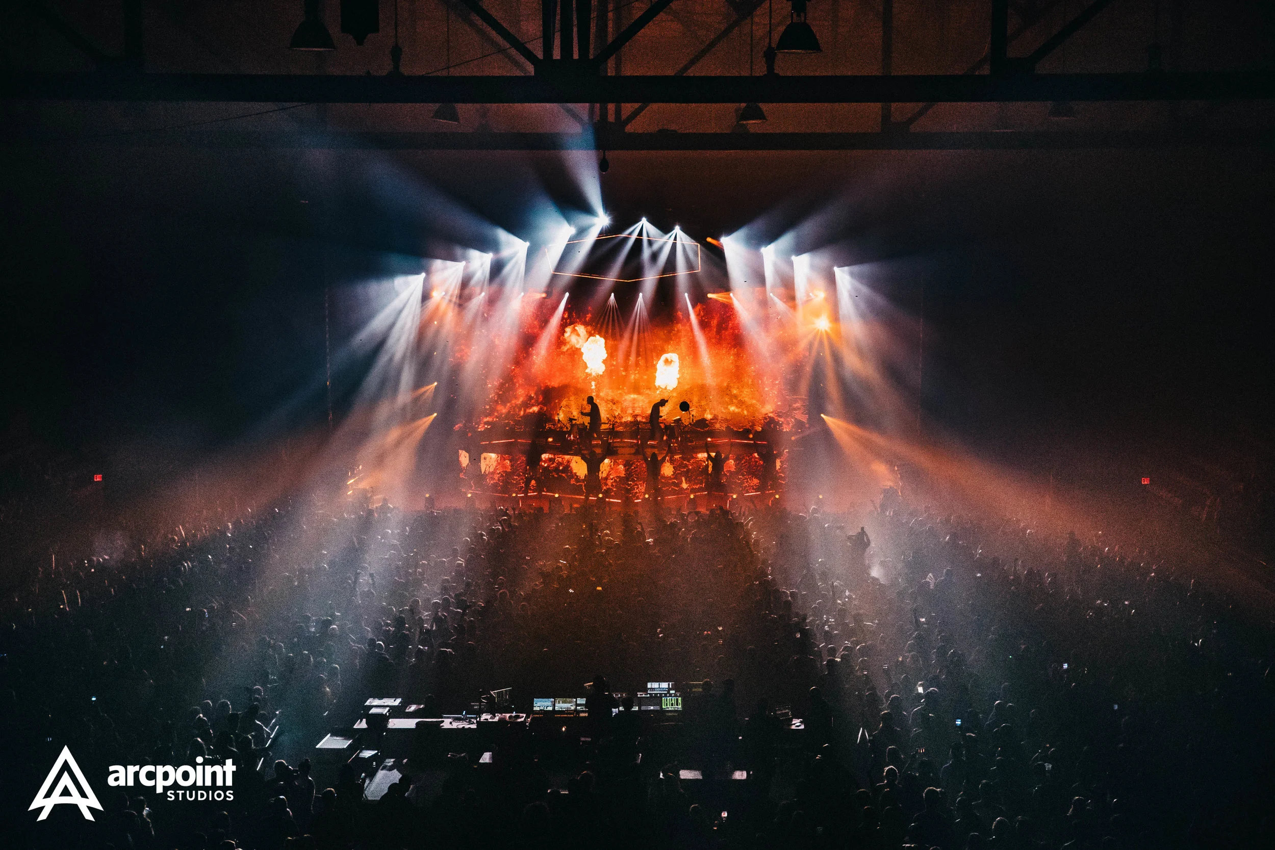 A large crowd inside a dark concert hall watching a live music performance with bright white and orange stage lights and a fiery fire display on the screen behind the performers.