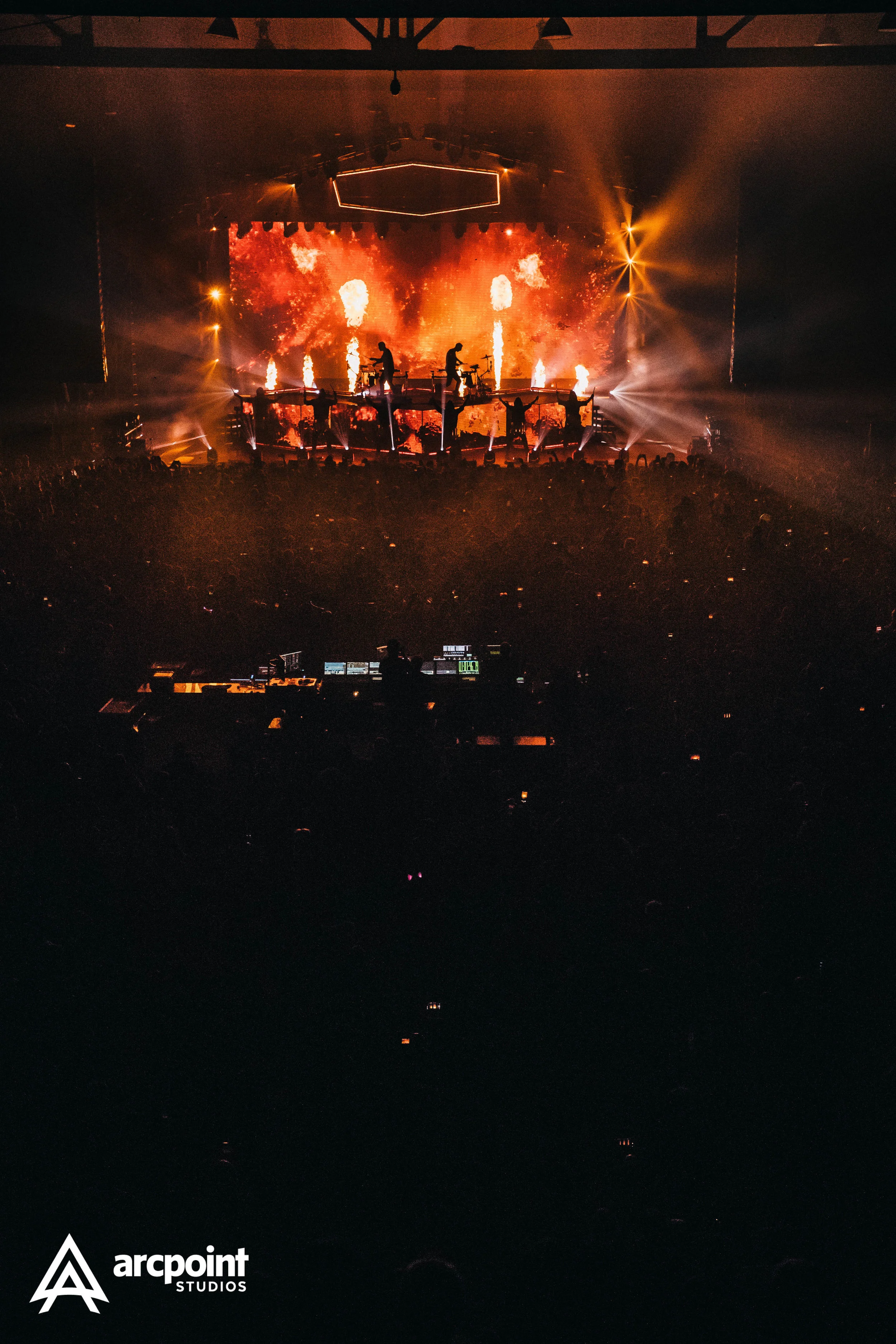 Stage with band performing under fiery red and orange lights and pyrotechnics, audience in darkness, with Arcpint Studios logo in the bottom left corner.