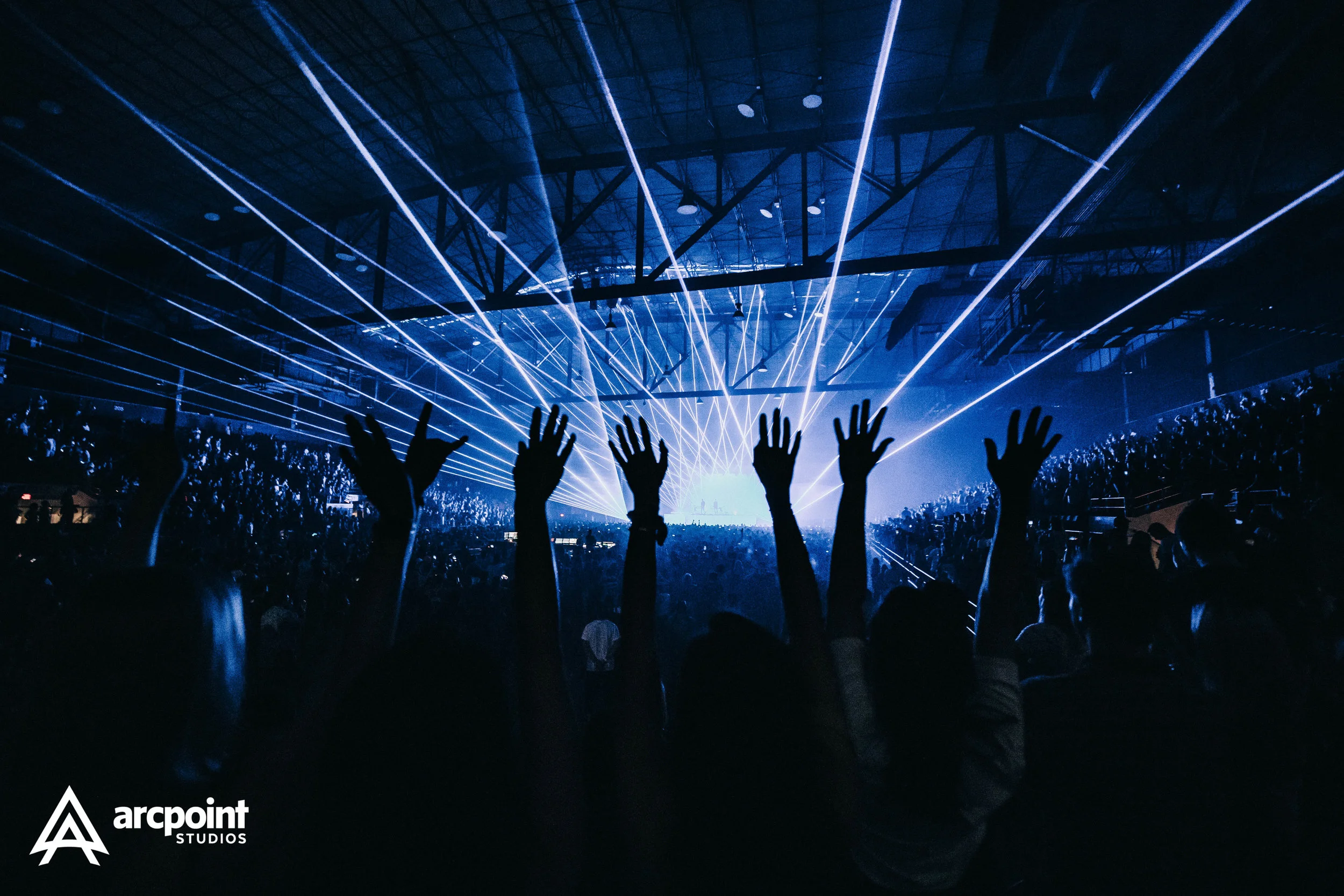 Crowd of people raising their hands at a concert or event with blue laser lights and stage in the background inside a large venue.
