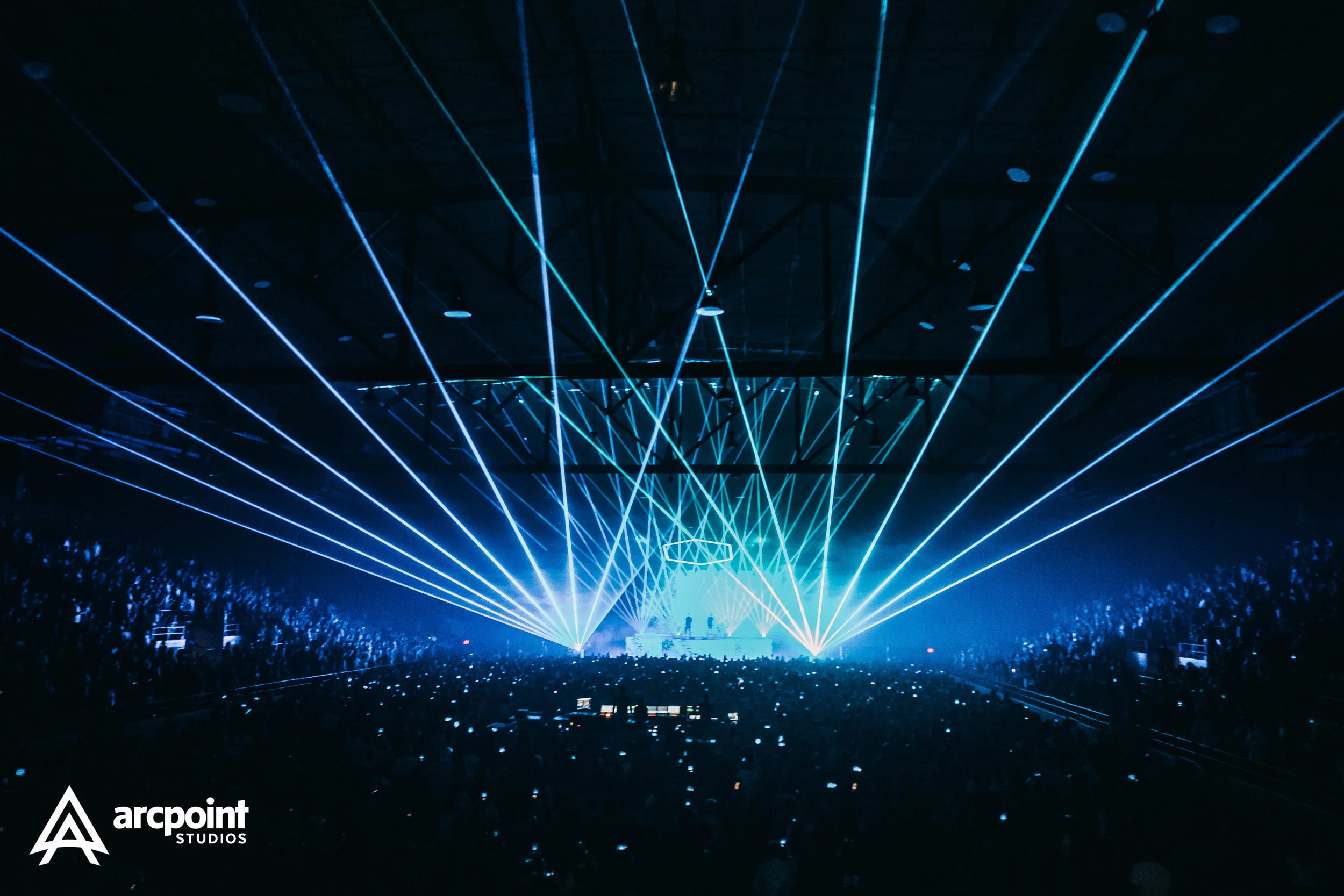 A concert or music event with blue laser lights projecting across the dark venue. The crowd is visible in the foreground, with many people holding up phones. The logo 'arcpoint STUDIOS' is in the bottom left corner.