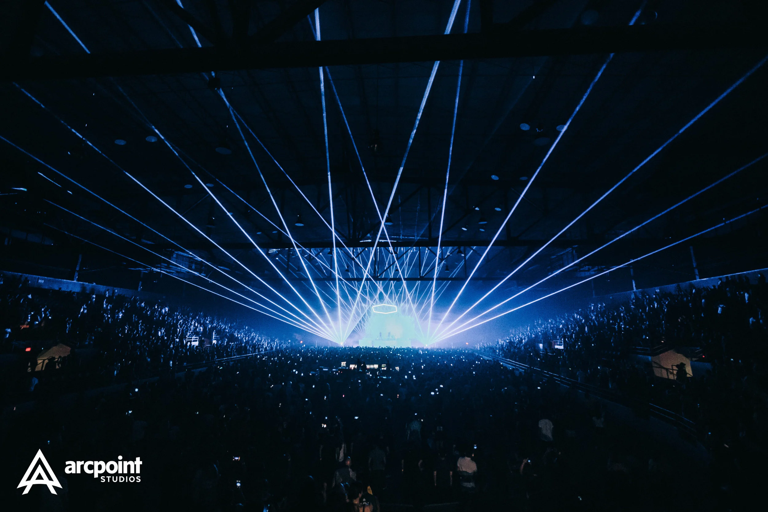 A large indoor concert with blue laser lights projecting from the stage into the air, and an audience watching. The venue is filled with people and the concert is broadcasting a lively atmosphere.