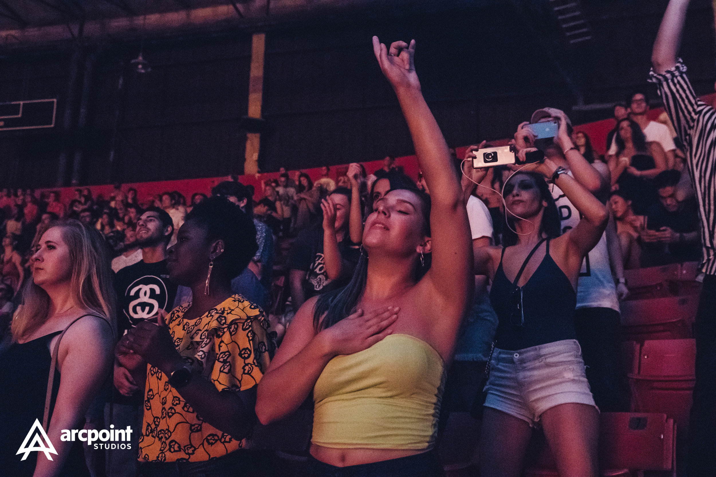 People in an indoor concert or event are standing closely together, some with their eyes closed, one woman with her hand over her chest and another with a hand raised, while others are recording or taking photos with their smartphones.