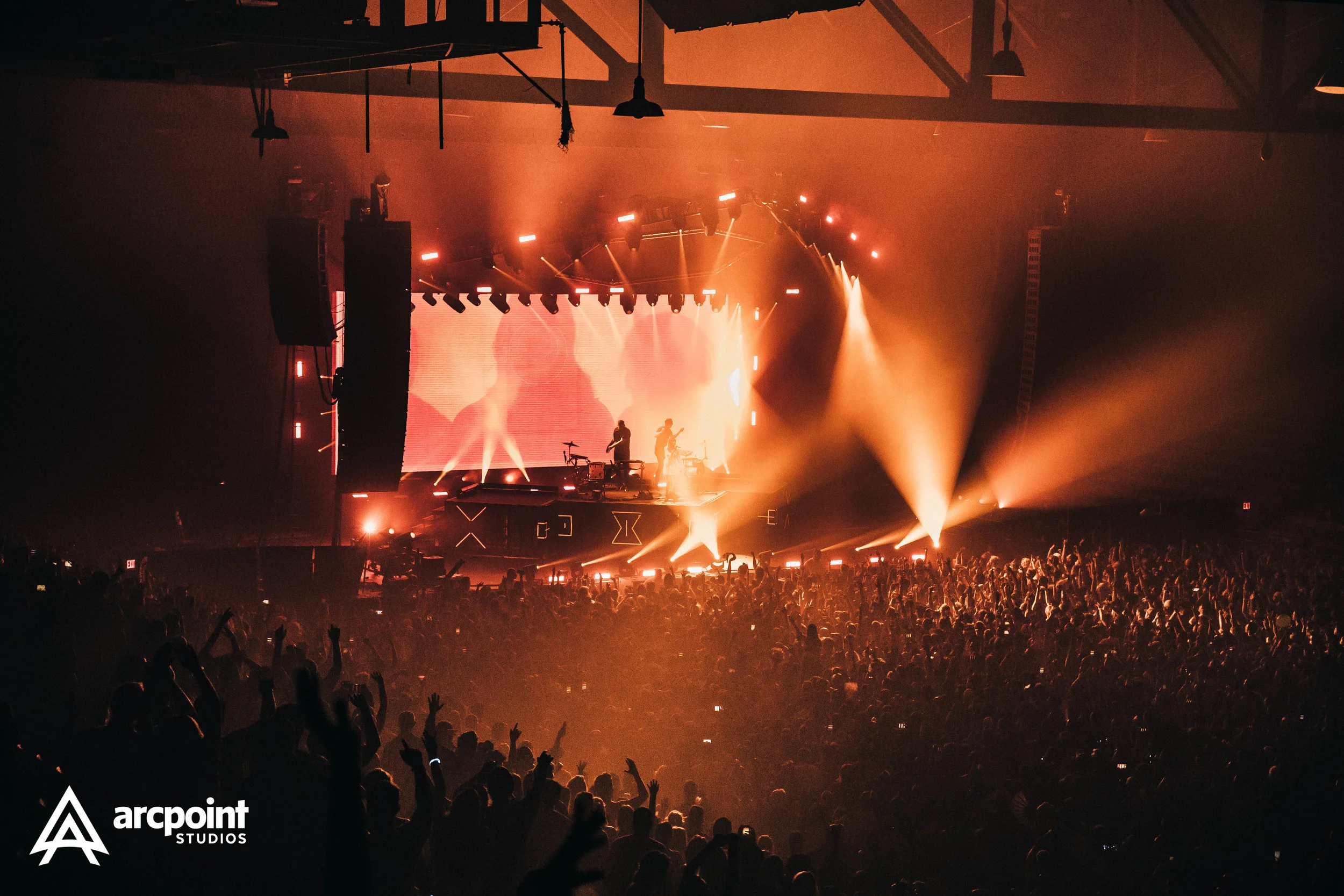 A concert stage illuminated with warm orange lighting, with a large crowd of people raising their hands in front. Performers are on stage, and a big digital screen displays a silhouette. The venue has high ceilings with visible beams and hanging ligh