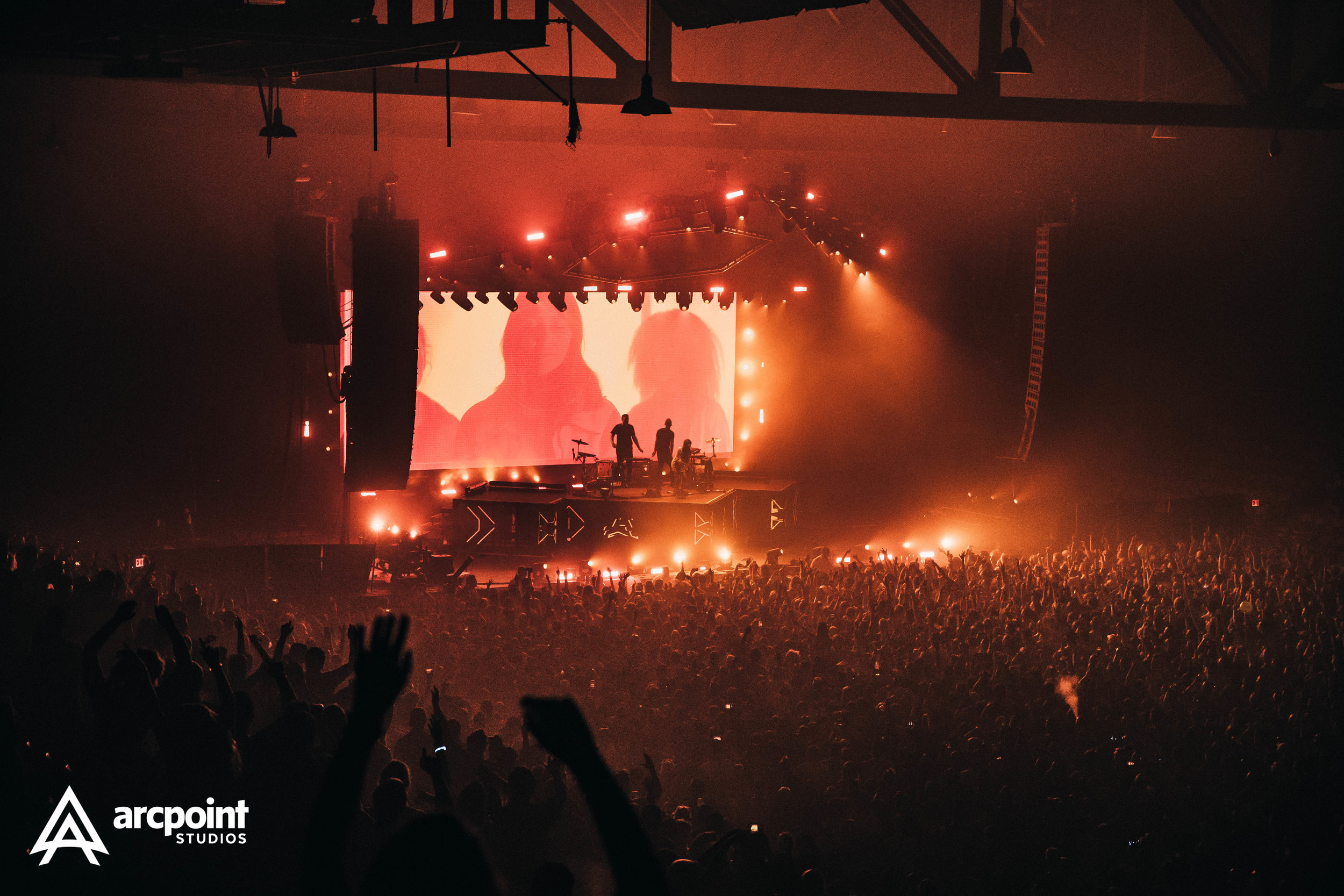 A concert scene with a large crowd in front of a stage bathed in orange and red lighting, with silhouettes of two performers, a large LED screen displaying abstract visuals, and various stage equipment.