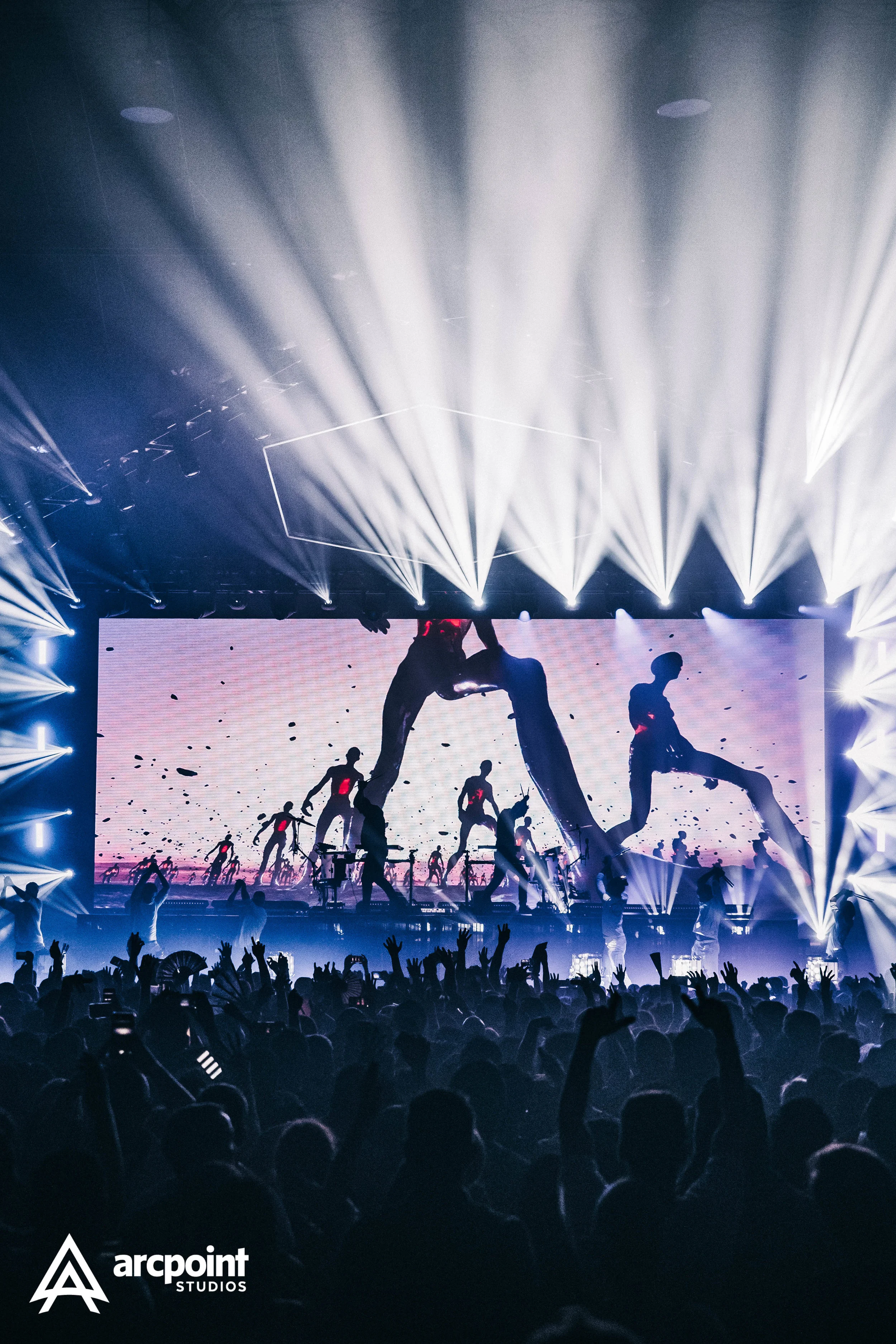 A large concert or dance performance with a stage featuring a digital screen showing dancers in silhouette. Bright white stage lights are shining from above onto the stage and the crowd in front is raising their hands. The bottom left corner shows th