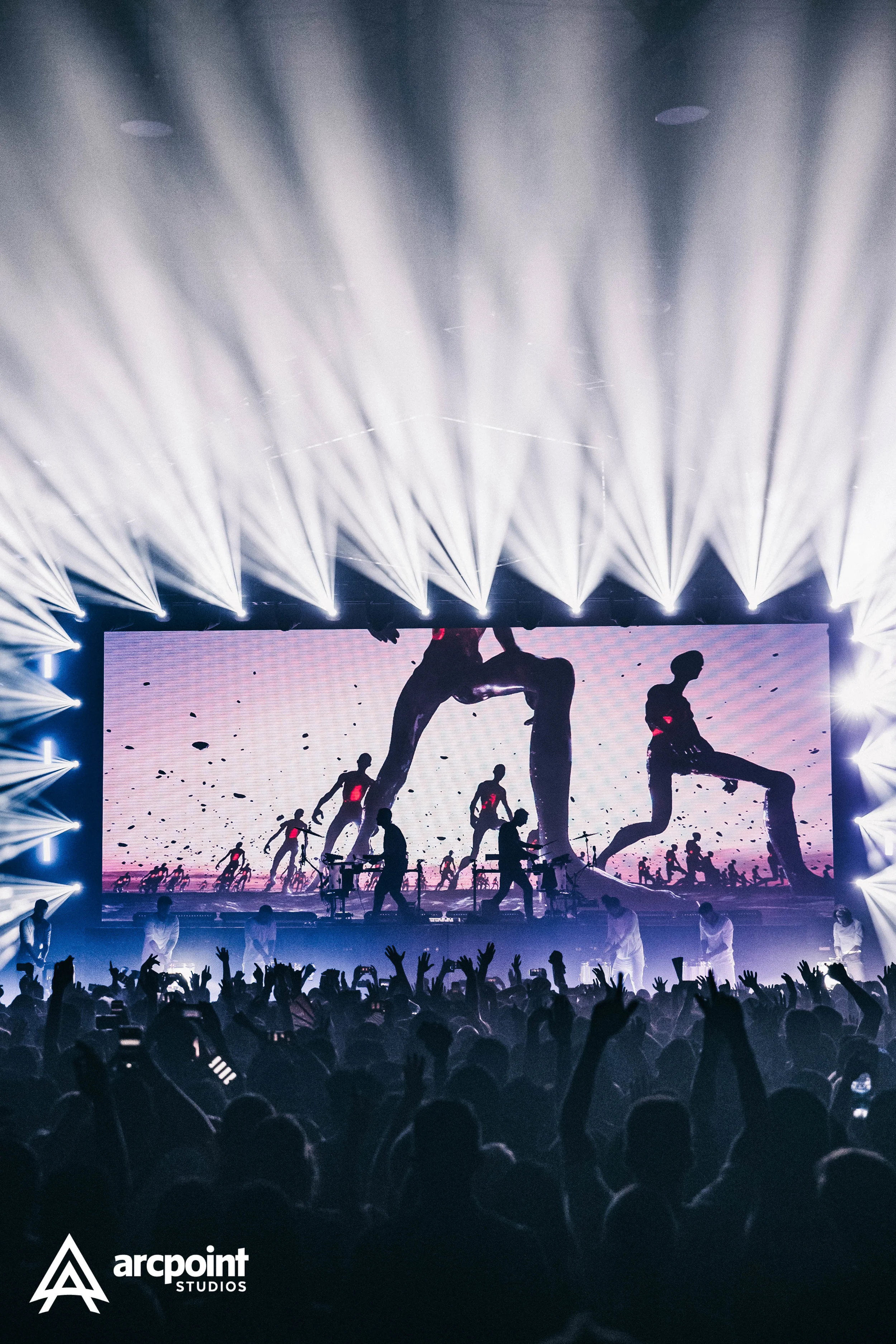 A concert or large event with a crowd in front of a stage, bright stage lights, and a large screen displaying silhouettes of dancers performing. The stage includes performers and the background shows multimedia visuals.