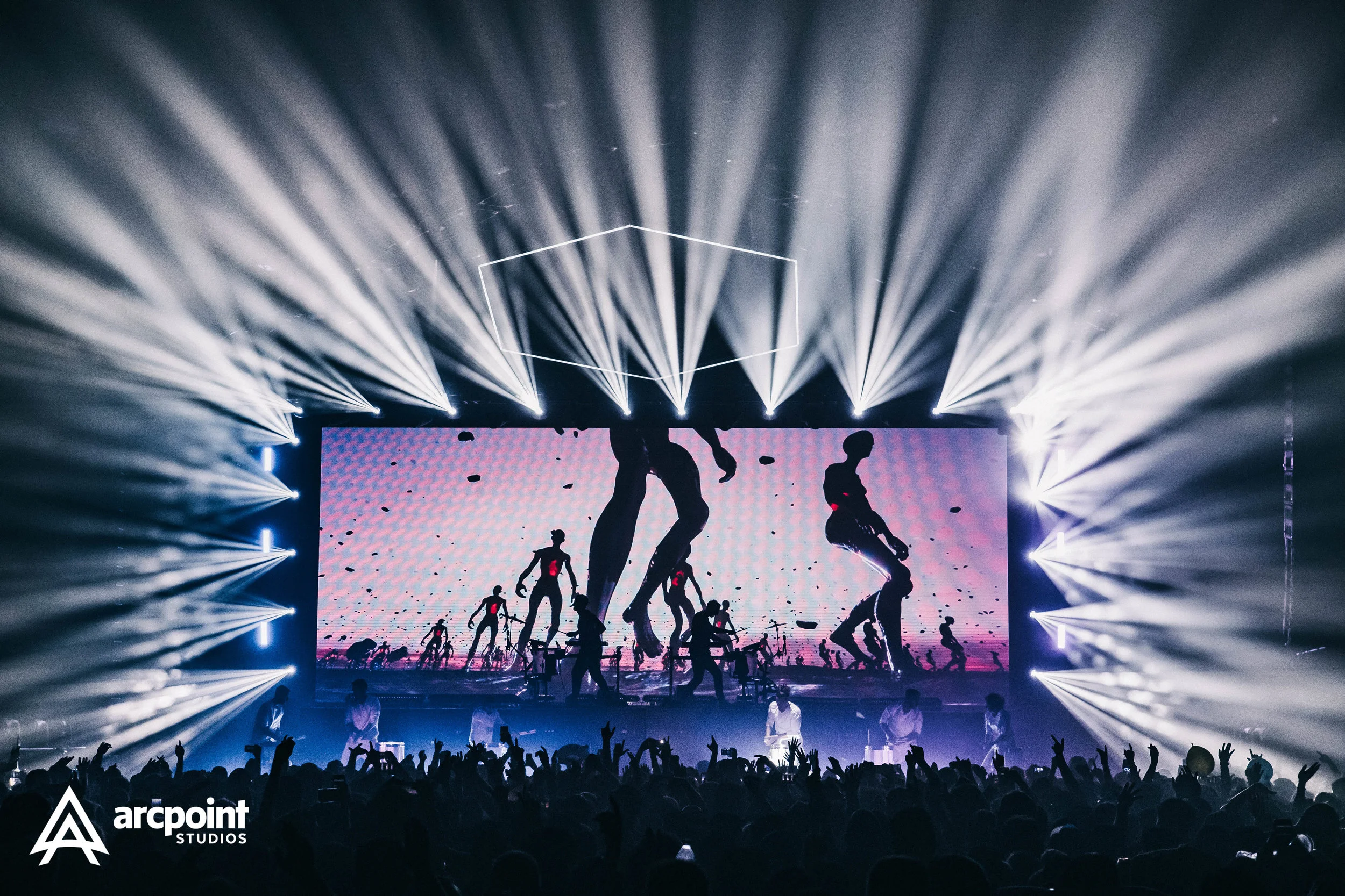 A concert or live event with bright stage lighting and a digital screen showing silhouettes of dancers or performers; large crowd with hands raised in the foreground; 'arcpoint STUDIOS' logo in bottom left corner.