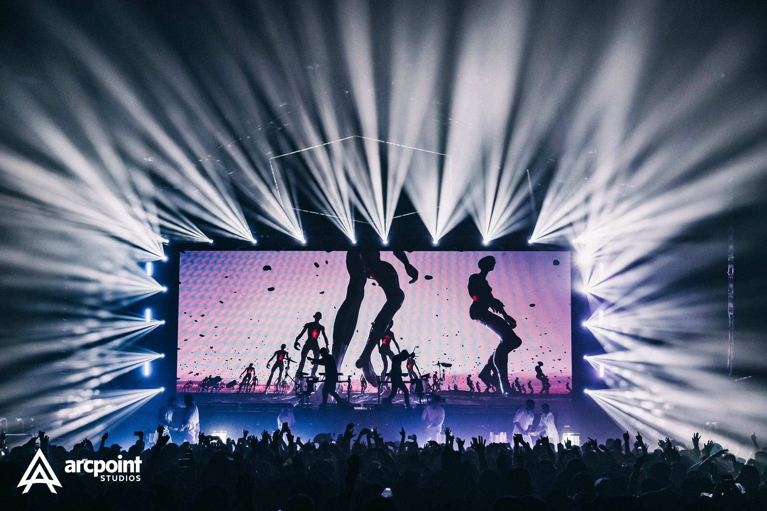 A concert scene with a large crowd, stage with performers, and a bright display screen showing silhouettes of people dancing. Light beams radiate above the stage.