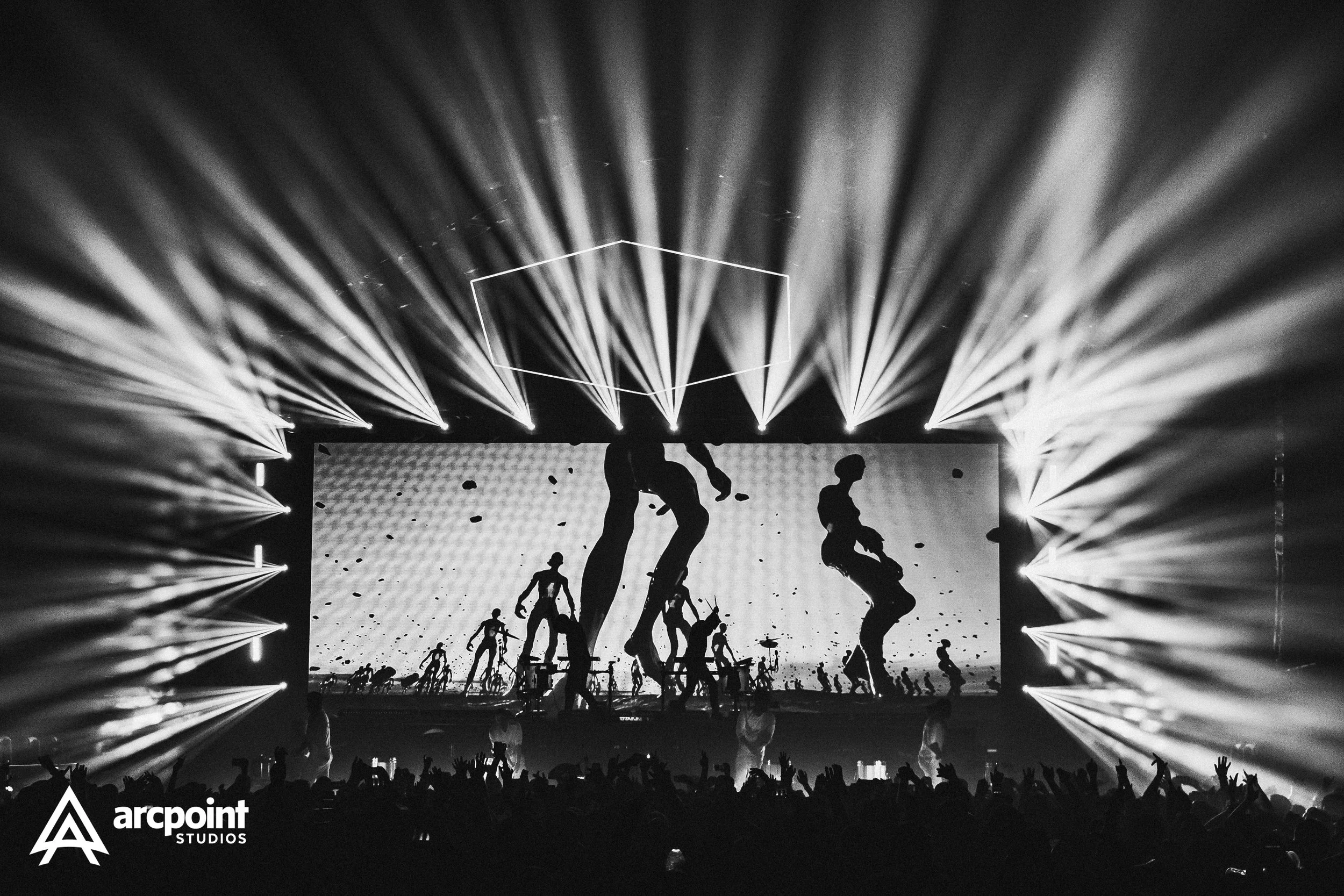 Black and white photo of a concert stage with bright stage lights and a large screen displaying silhouetted figures of people performing or dancing, with a crowd of people in front.