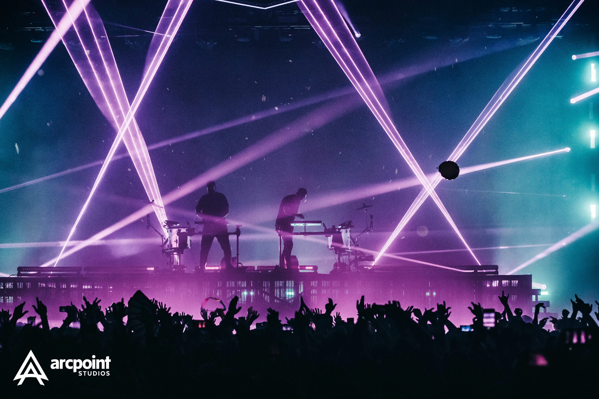 Silhouettes of two musicians performing on stage with colorful laser lights and an audience in front, at a concert.