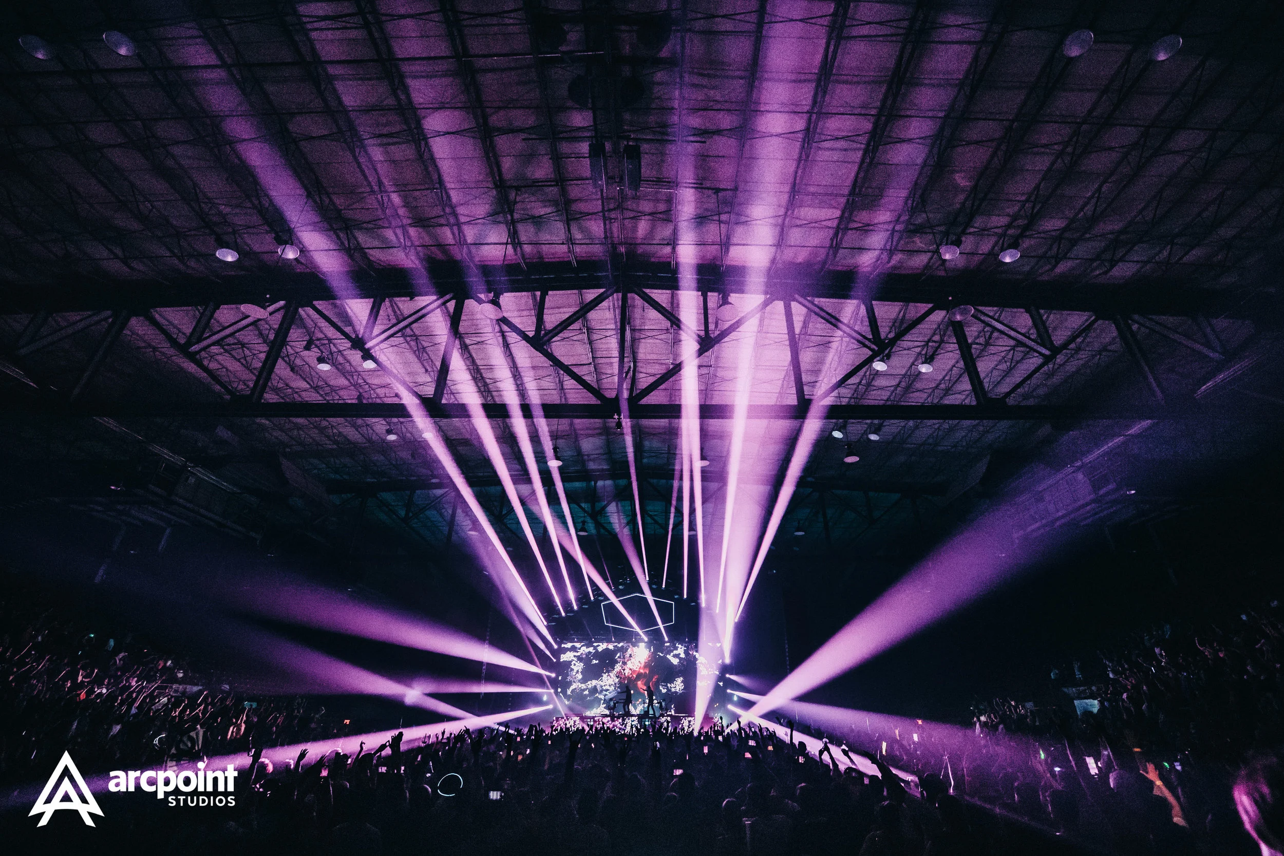 A concert with purple stage lights, a crowd of people with raised hands, and a DJ or performer on stage, inside a large indoor venue with a high ceiling.