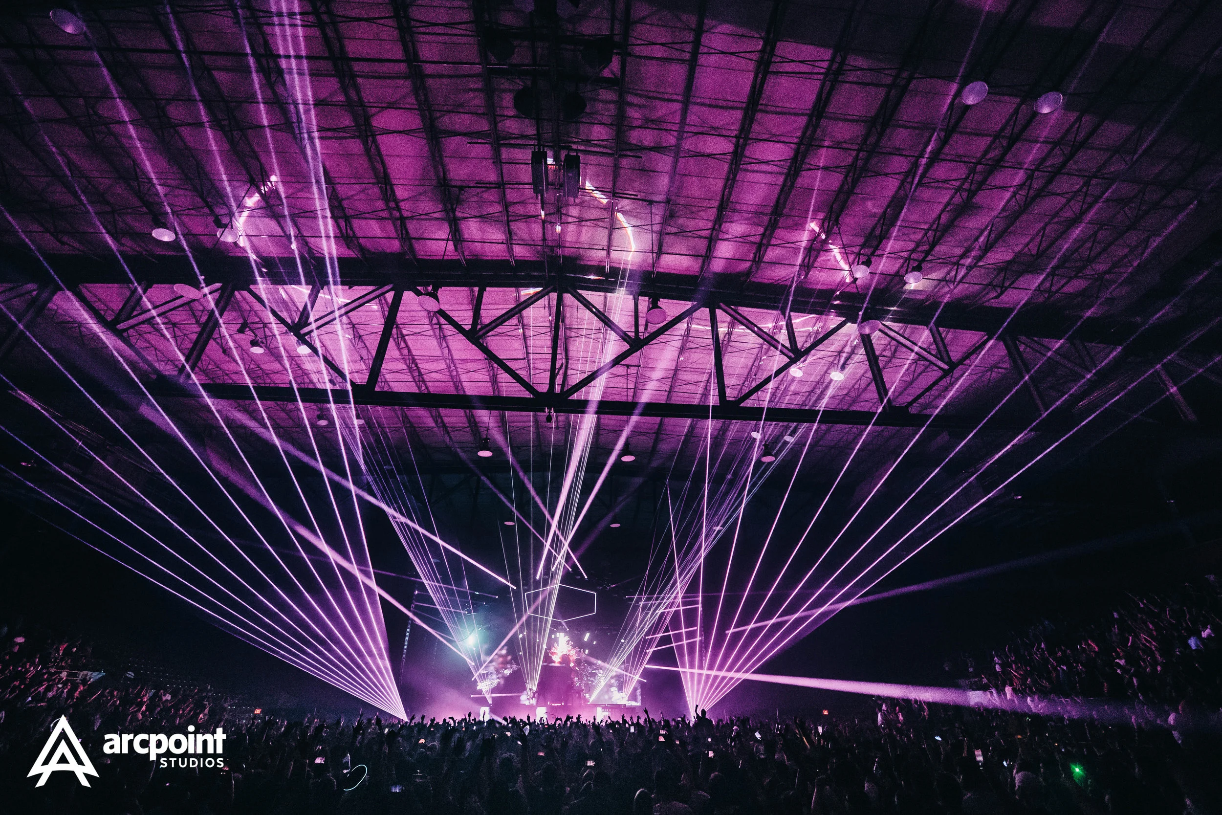 A concert venue with a large crowd, bright purple laser lights, and a stage setup, with the Arcpont Studios logo in the bottom left corner.