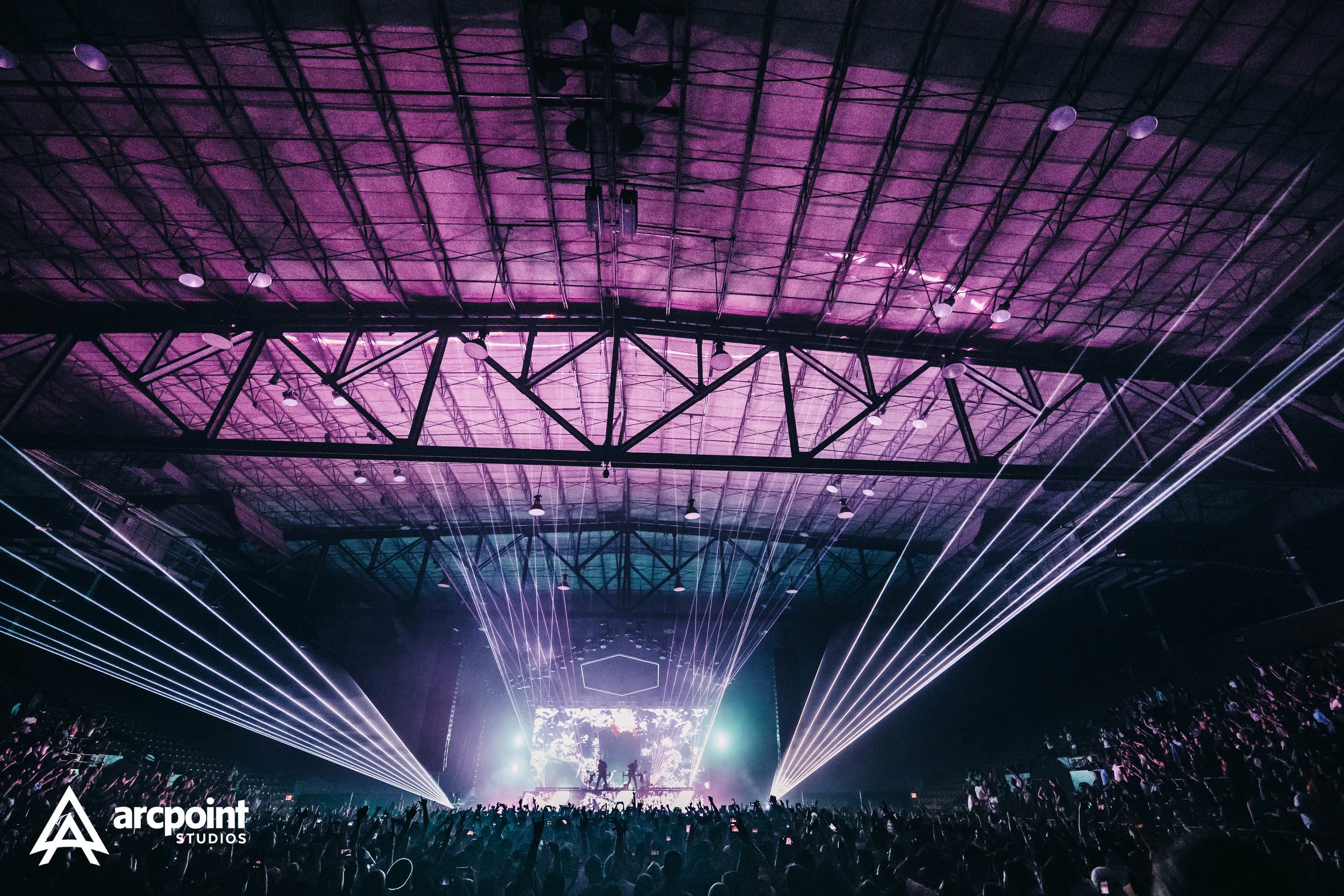 An indoor concert with a large crowd, stage lighting, and laser lights in a spacious venue with a high ceiling.