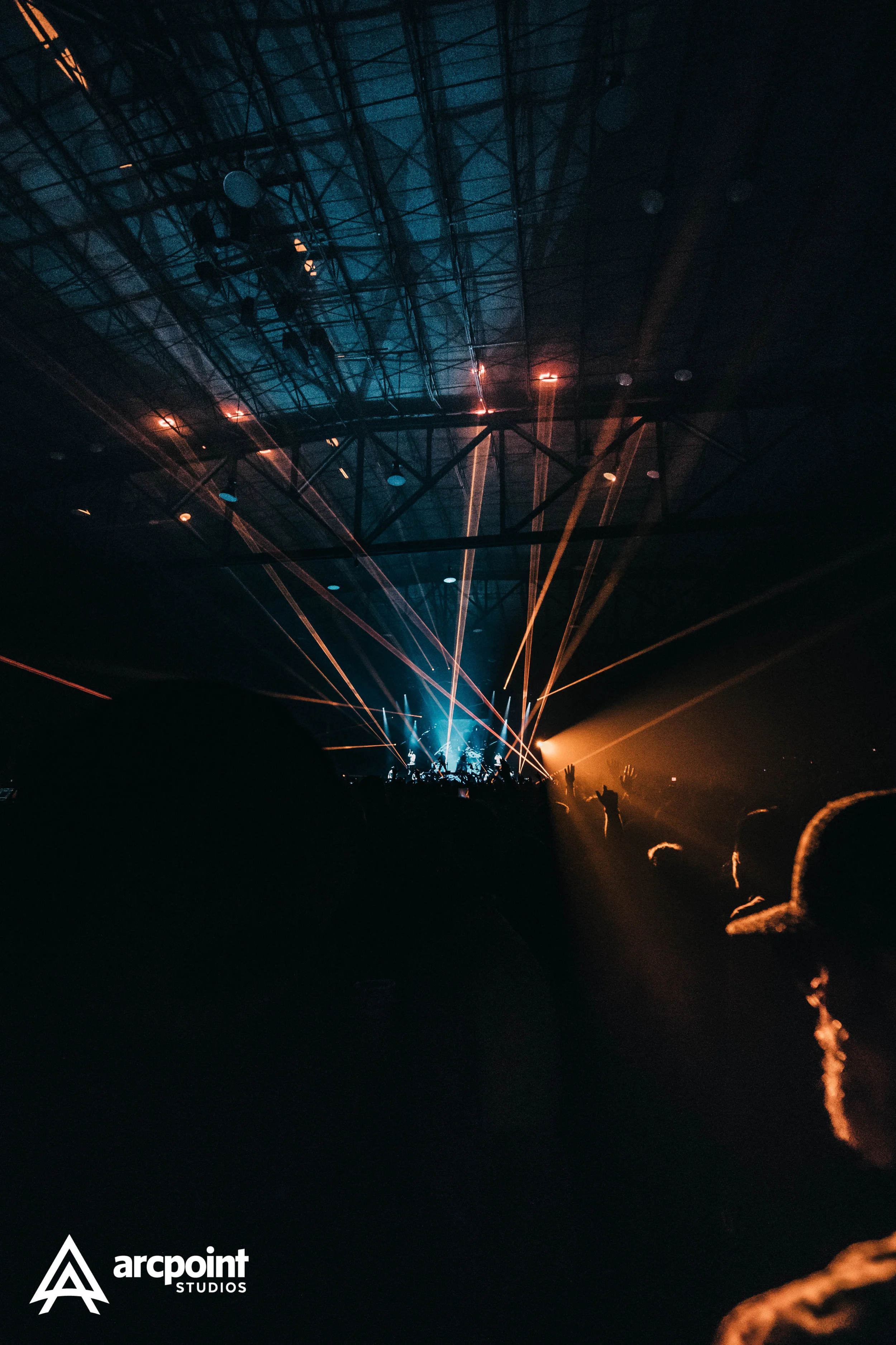 Concert scene with colorful laser lights and audience members with raised hands in a large indoor venue.