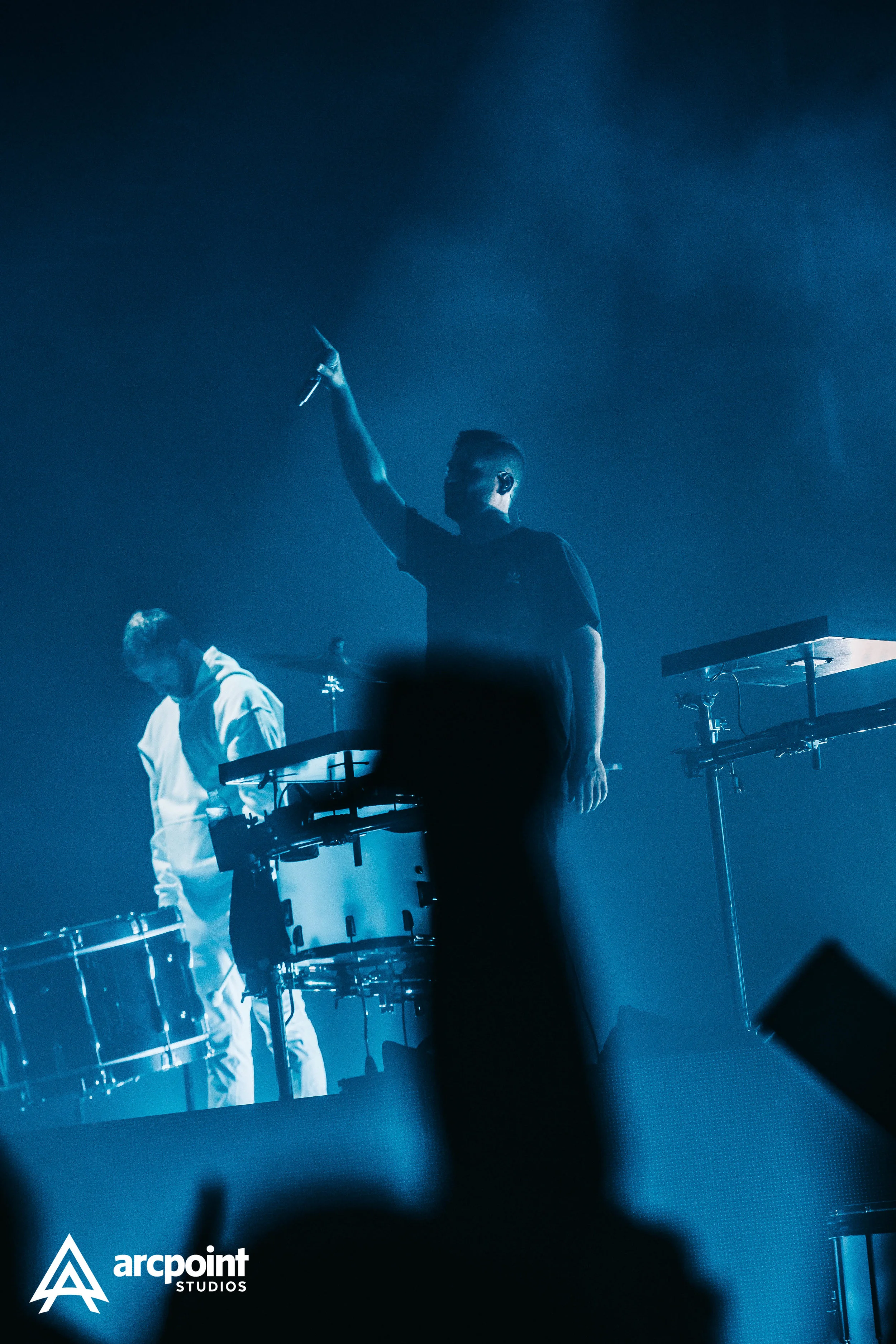 Two performers on stage with electronic drums, one raising their arm, the scene illuminated with blue lighting, logo of arcpoint studios in the bottom left corner.