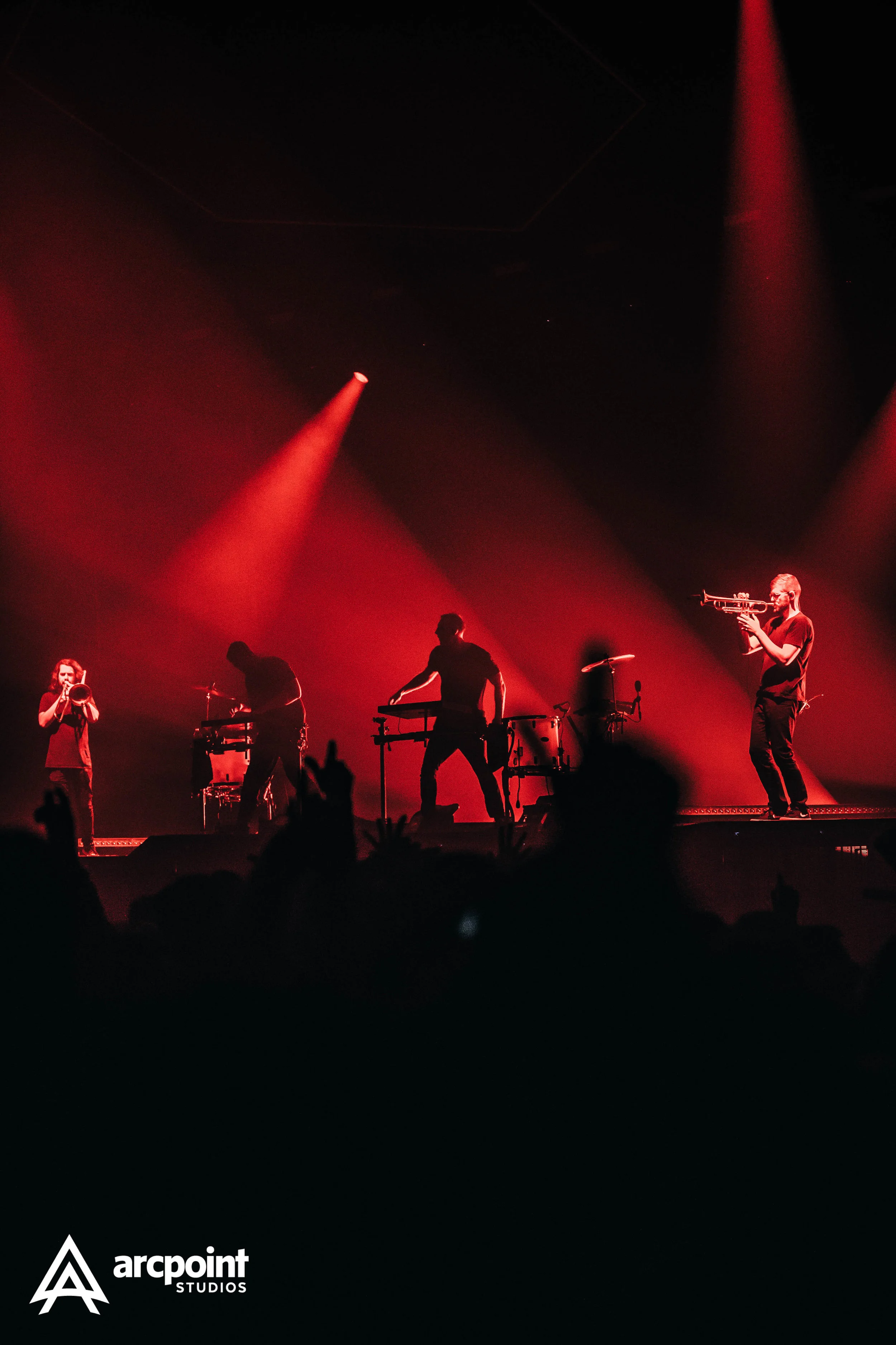 A live music performance with four musicians on stage, silhouettes against red lighting, one playing a trumpet, another playing a drum, and two others with electronic instruments.