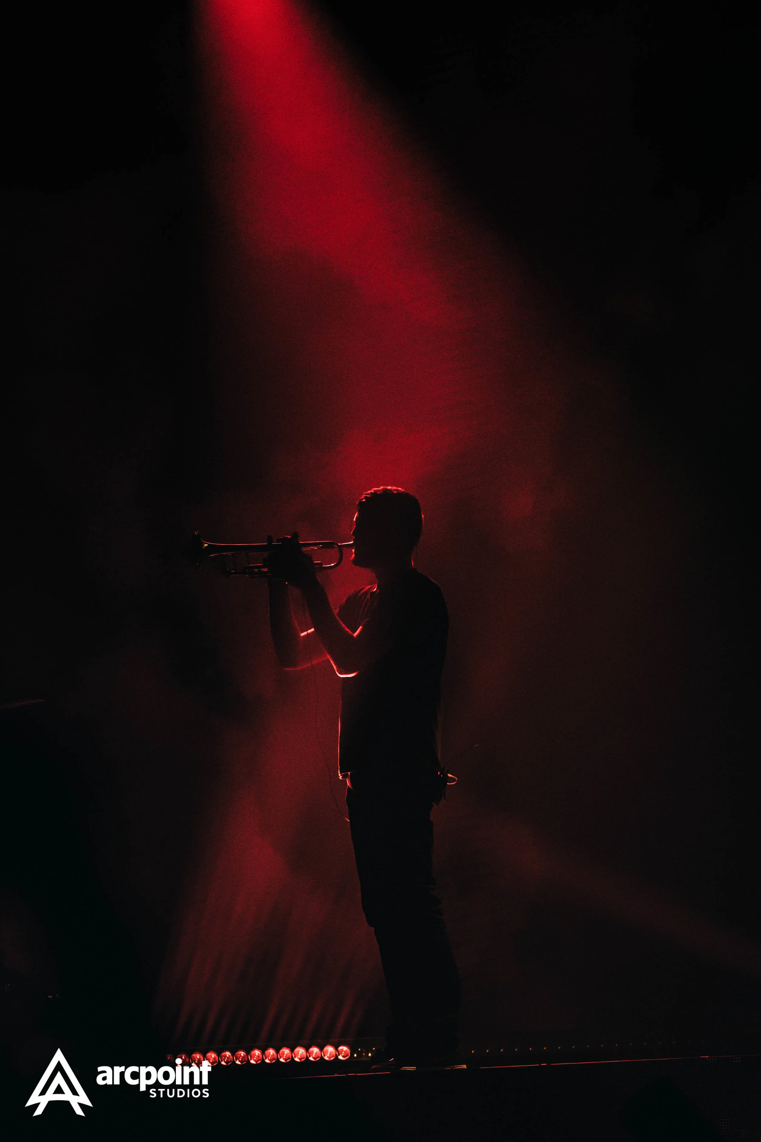 Silhouette of a person playing the trumpet on stage illuminated by red stage lights with the 'arcpoint STUDIOS' logo in the bottom left corner.