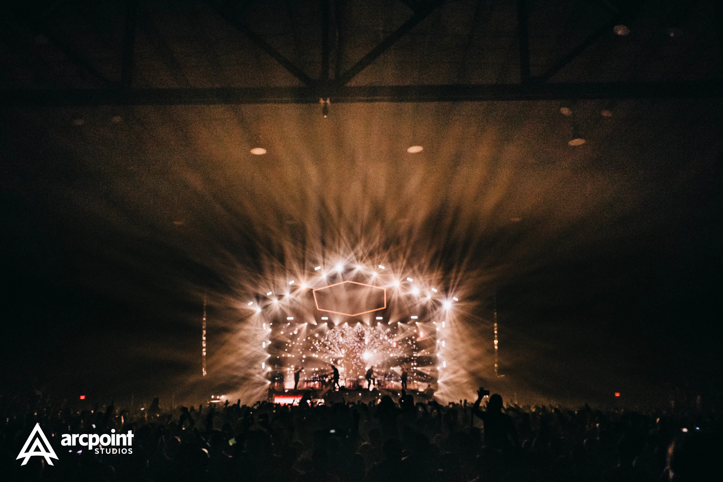 A concert scene with bright lights and a large crowd, featuring a stage with electronic equipment and performers, with smoke and dynamic lighting effects.