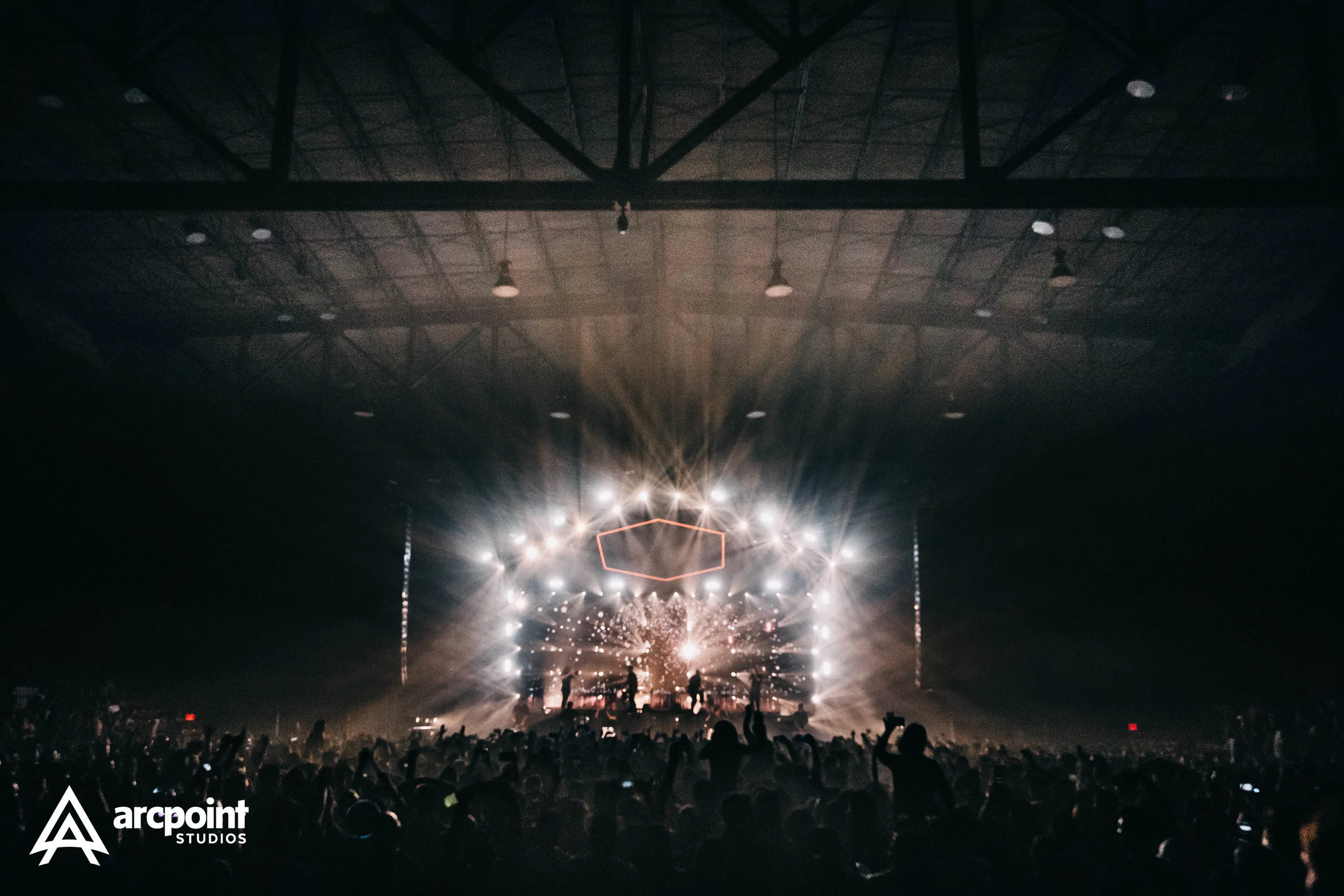 Large indoor concert arena filled with a crowd of people watching a stage with bright lights and sparkles. The logo of arcpoint STUDIOS is in the lower left corner.