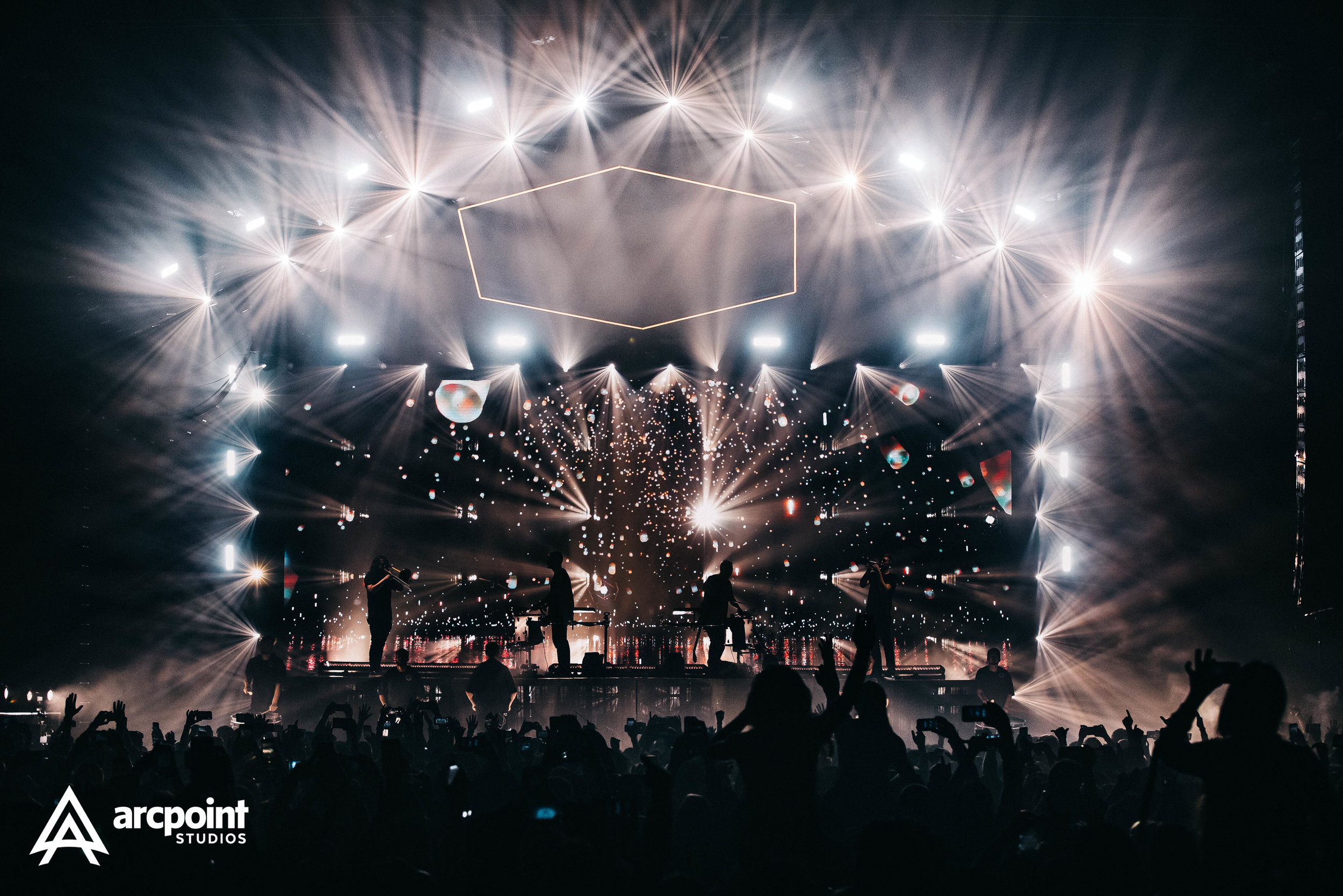 A concert stage with bright lights and graphics, silhouettes of musicians and a crowd watching.