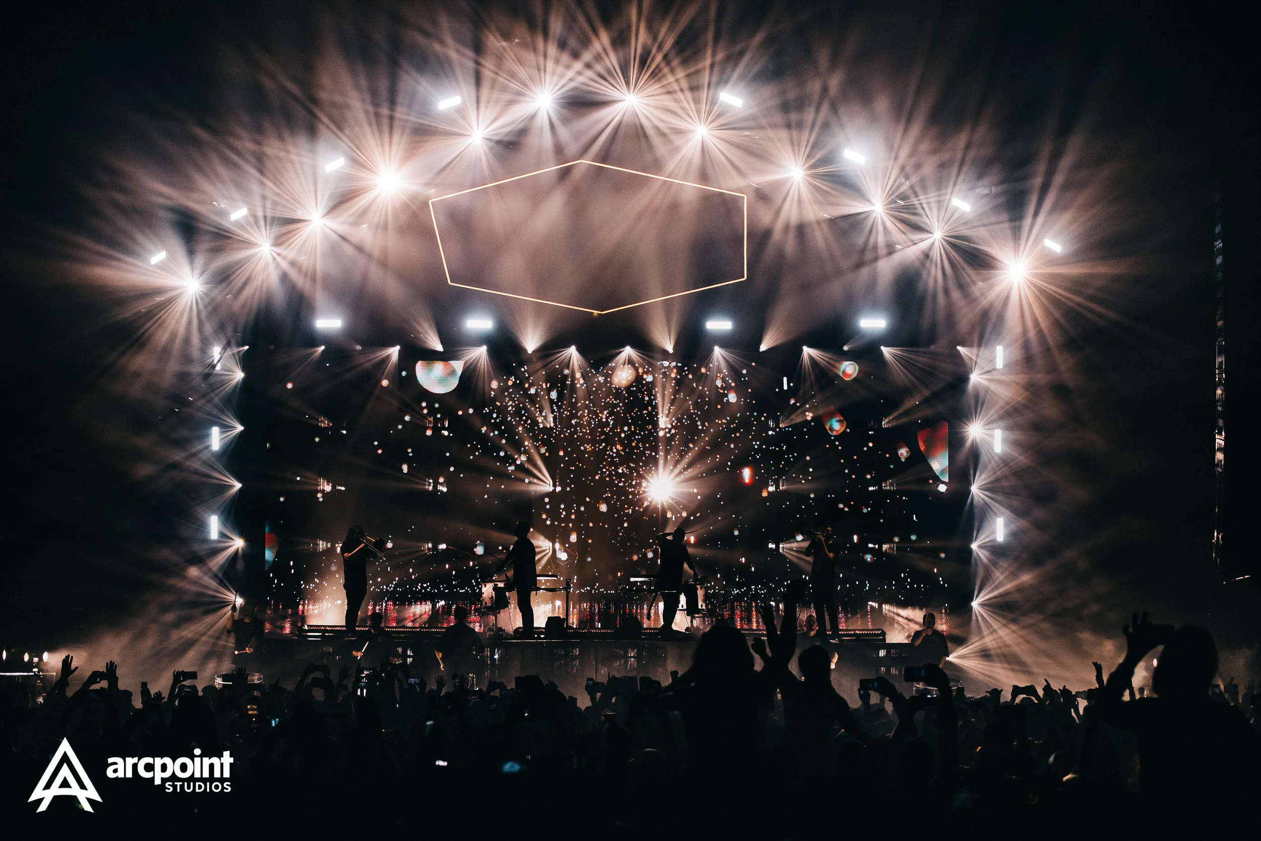 A live concert with a band performing on stage, featuring elaborate lighting and visual effects. Silhouettes of audience members can be seen in the foreground, some holding up phones.