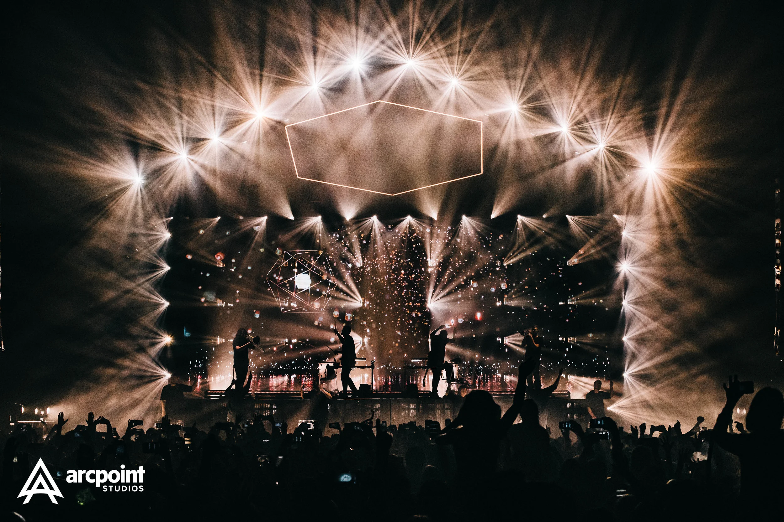 A concert stage with bright lights and geometric decorations, silhouettes of performers, and an audience capturing the moment with their phones.