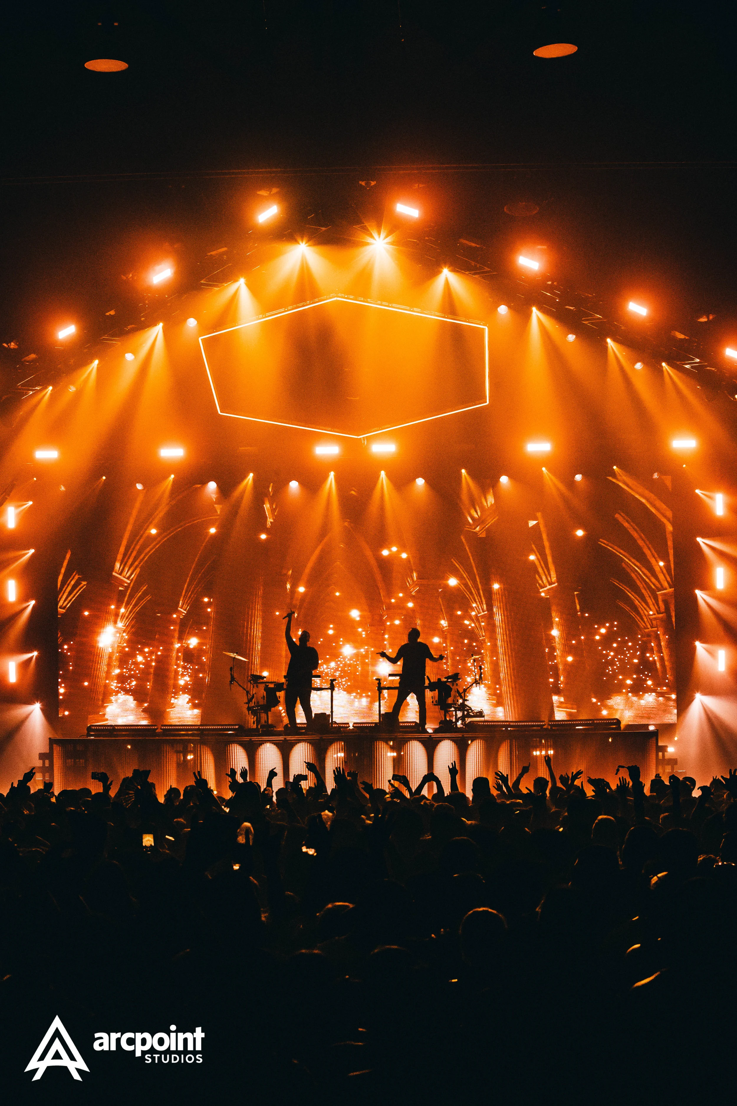 Concert scene with two silhouettes of DJs on stage, surrounded by bright orange and yellow stage lights, and an audience raising their hands in front of the stage. The logo for Arcpoint Studios is in the bottom left corner.