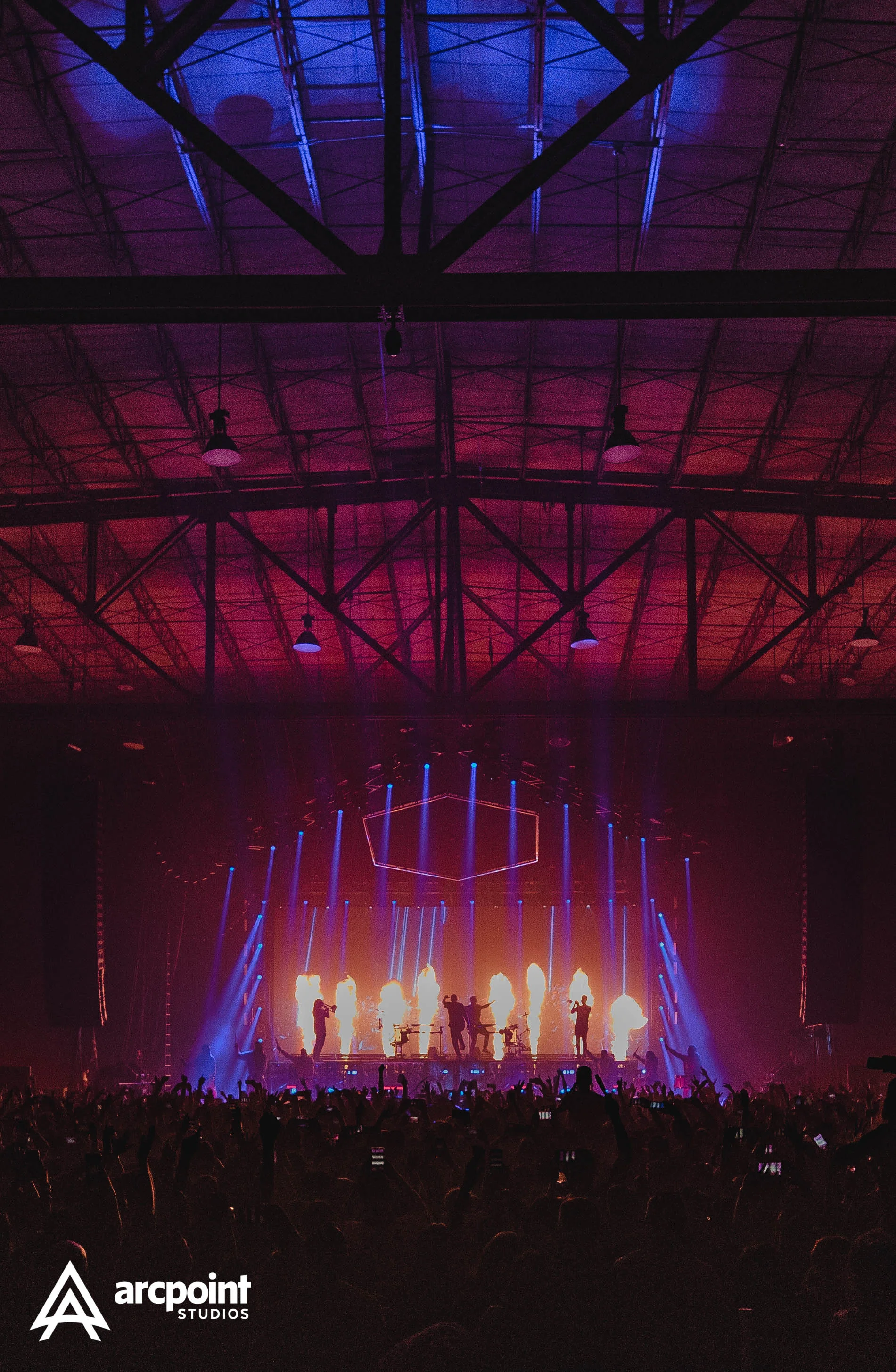 A concert or live performance with a stage illuminated by colorful lights and flames, showing silhouetted performers and a large audience in front.