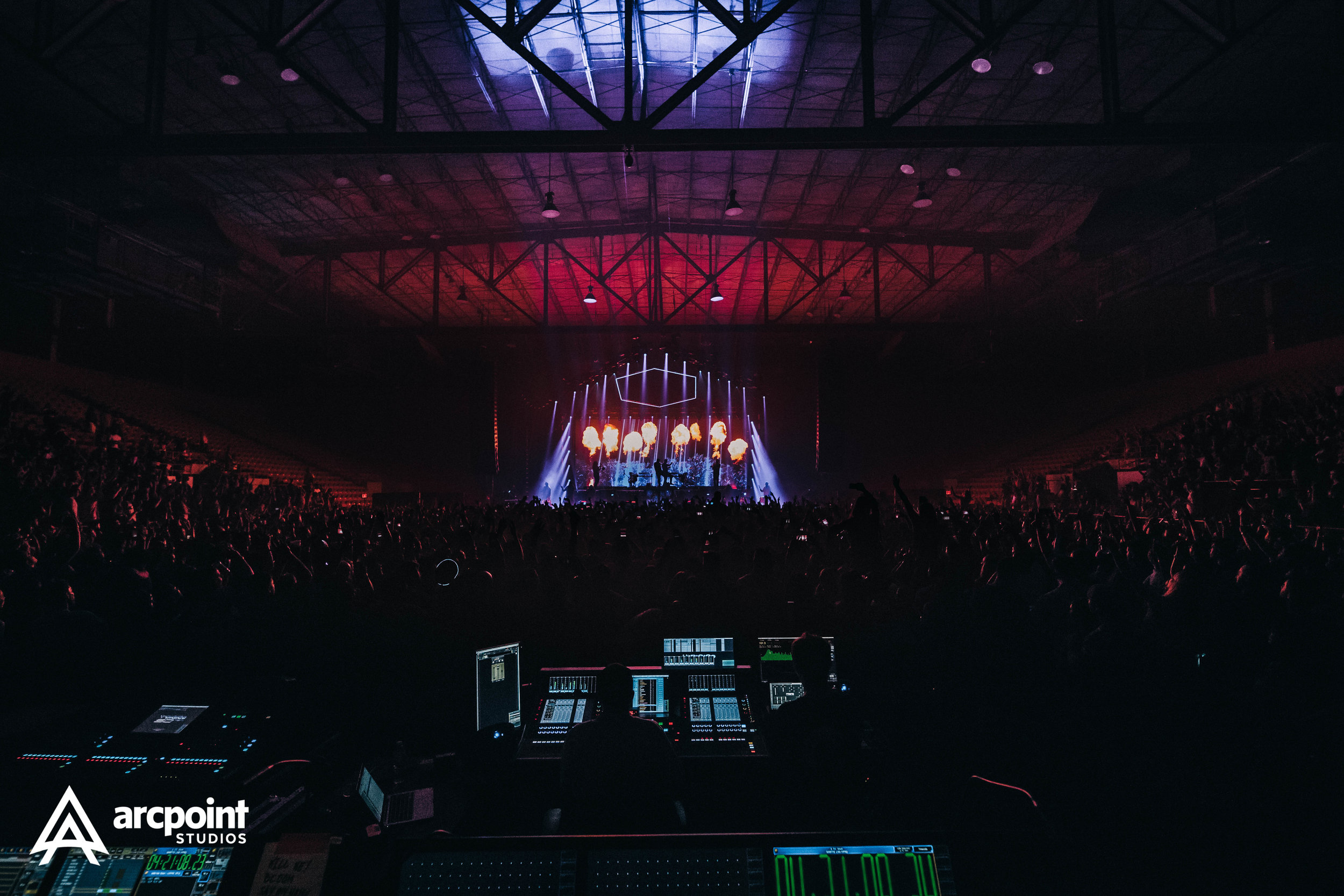 An indoor concert with a stage featuring vibrant lights and flame effects, viewed from the back with the audience and sound technicians in the foreground in a large arena.